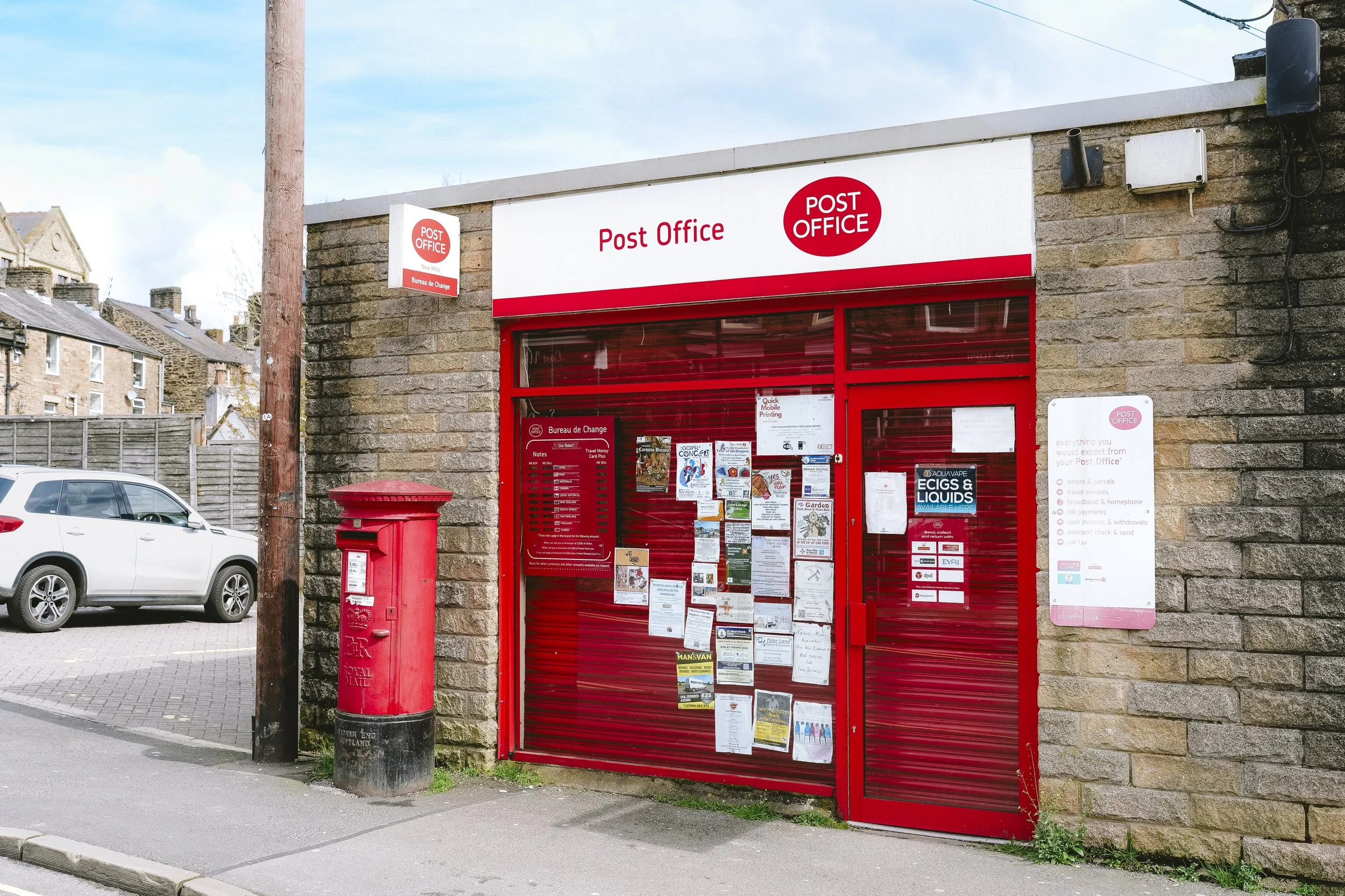 A small post office with a red door and mailbox, notices on the window, and signs indicating postal services. A car is parked nearby, and a utility pole is in front of the building.