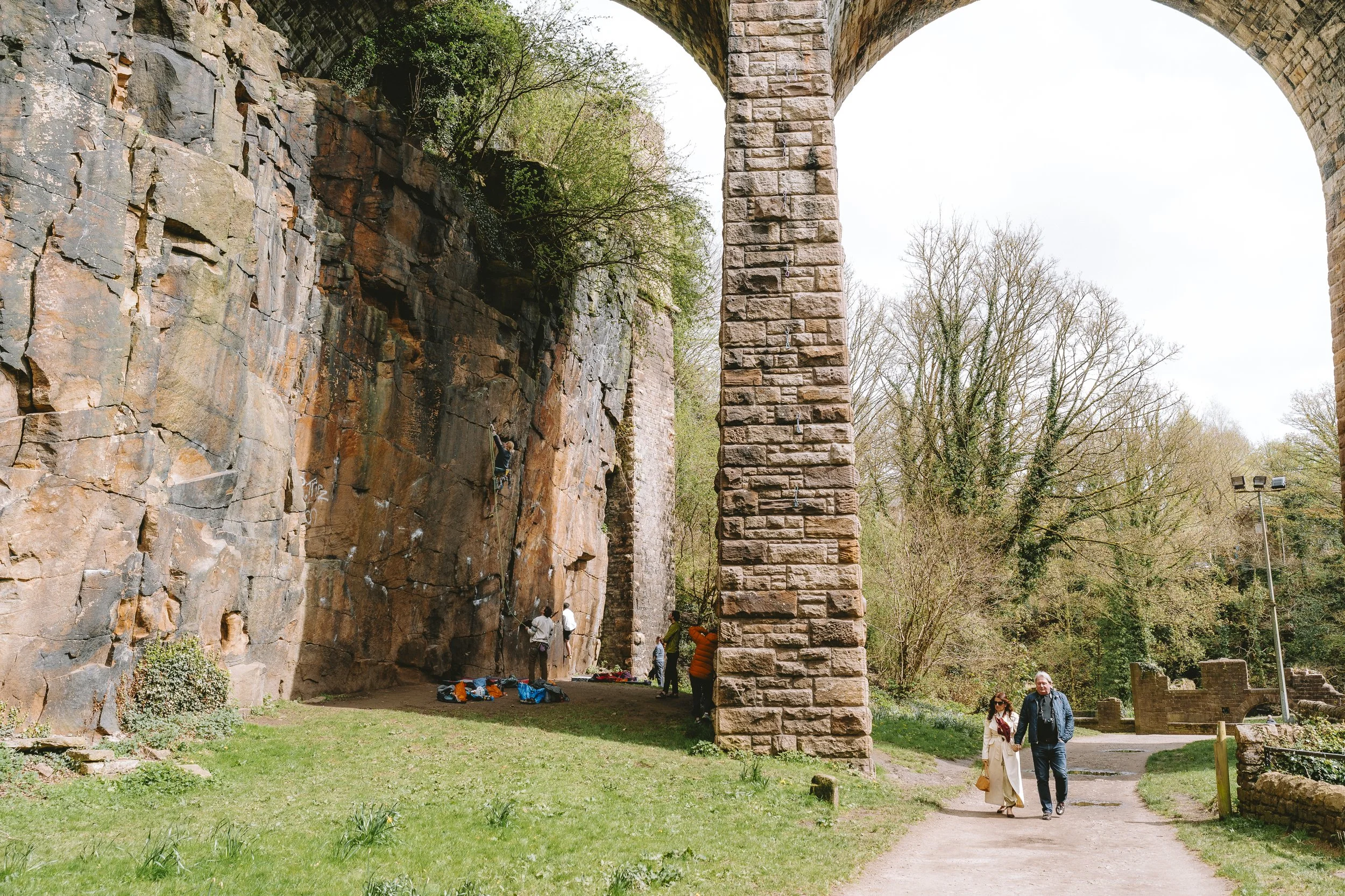 People walking near large stone and brick arch structures in a park with trees and grass.