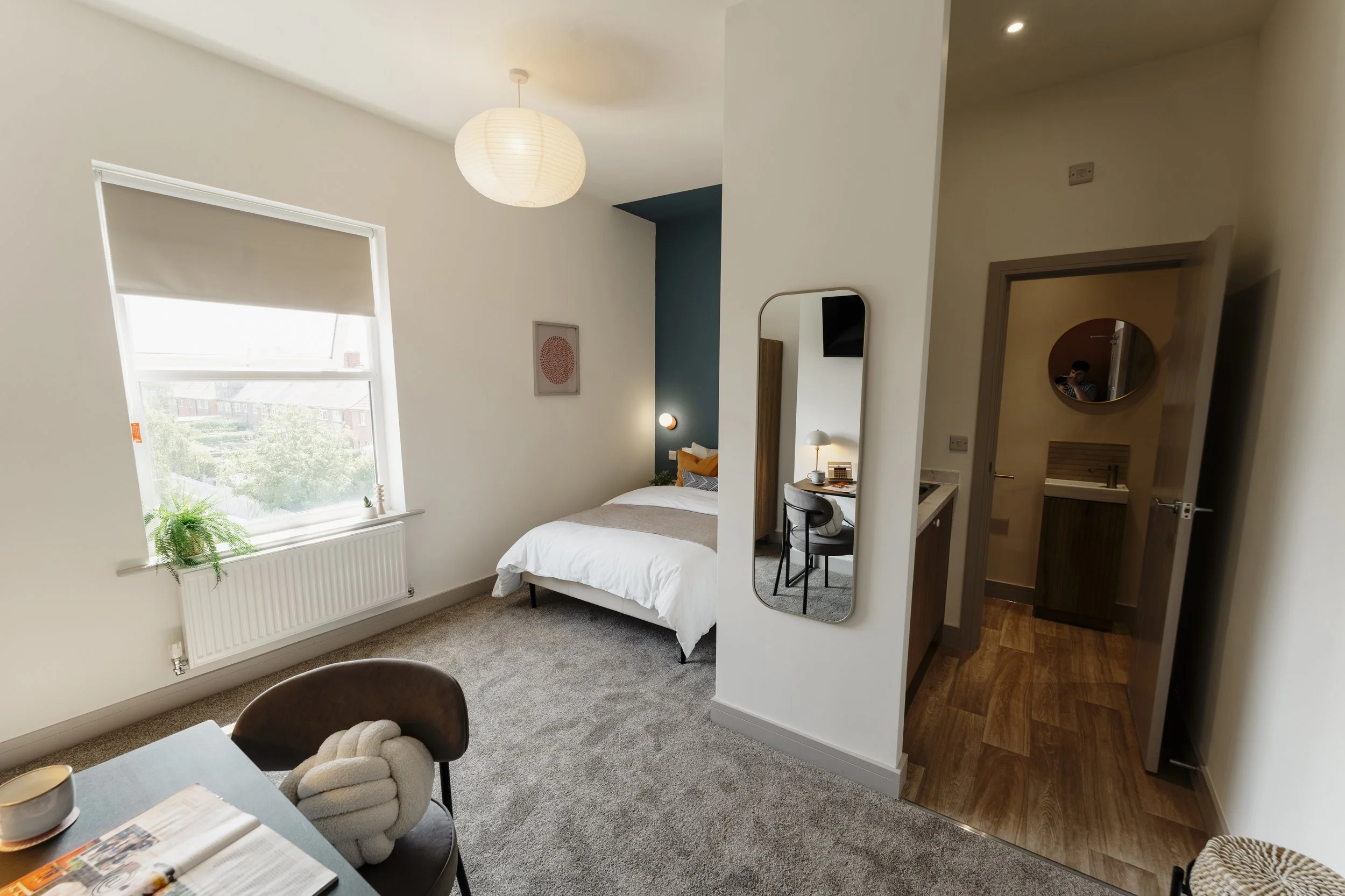A modern bedroom with a bed, a framed artwork on the wall, a large window with a white blind, a mirror on the wall, a bedside table with a lamp, and a partial view of a bathroom door. The room has beige carpeting and a pendant ceiling light.
