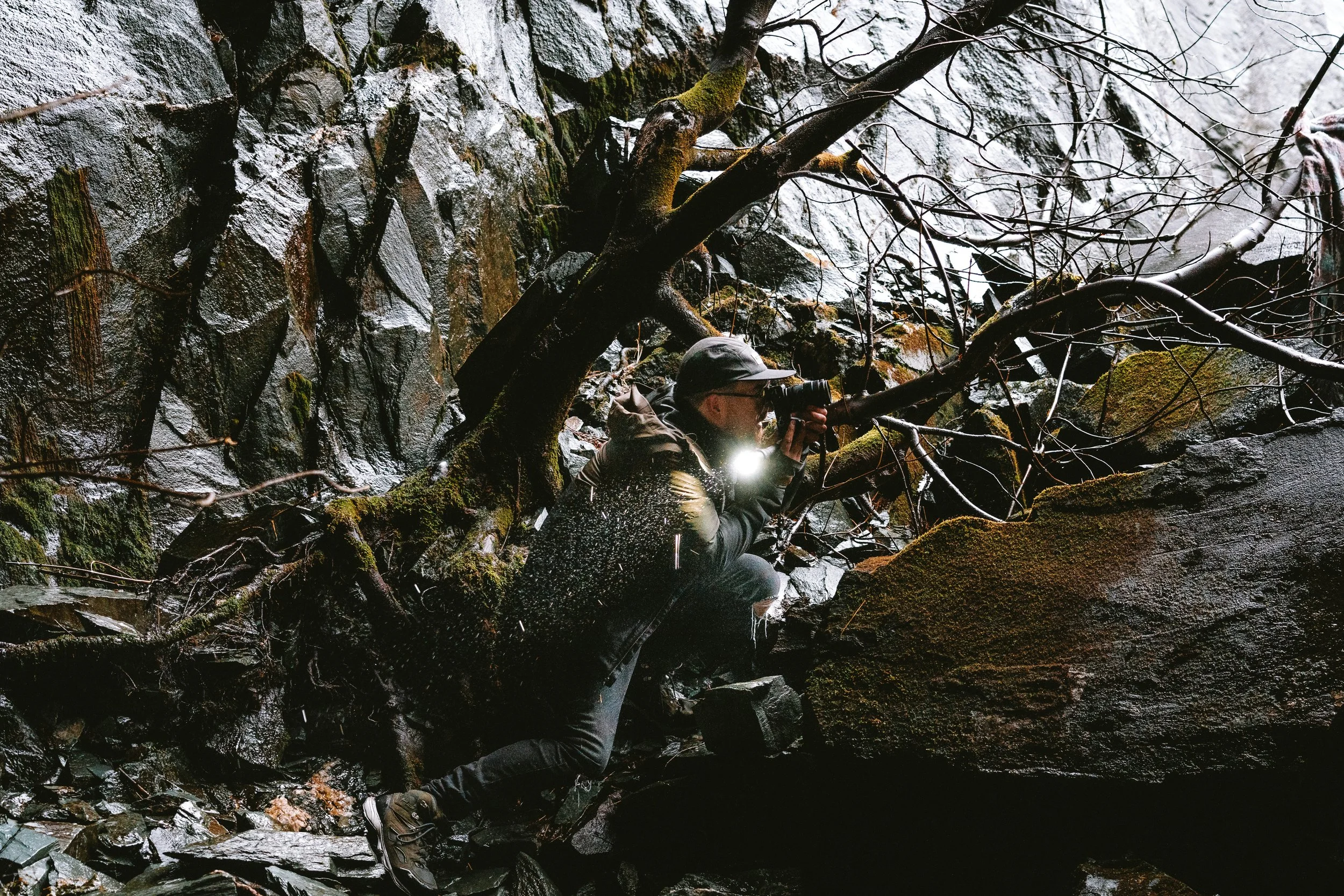 Person wearing outdoor gear with a headlamp, climbing through a rocky, wet ravine with trees and moss-covered rocks.