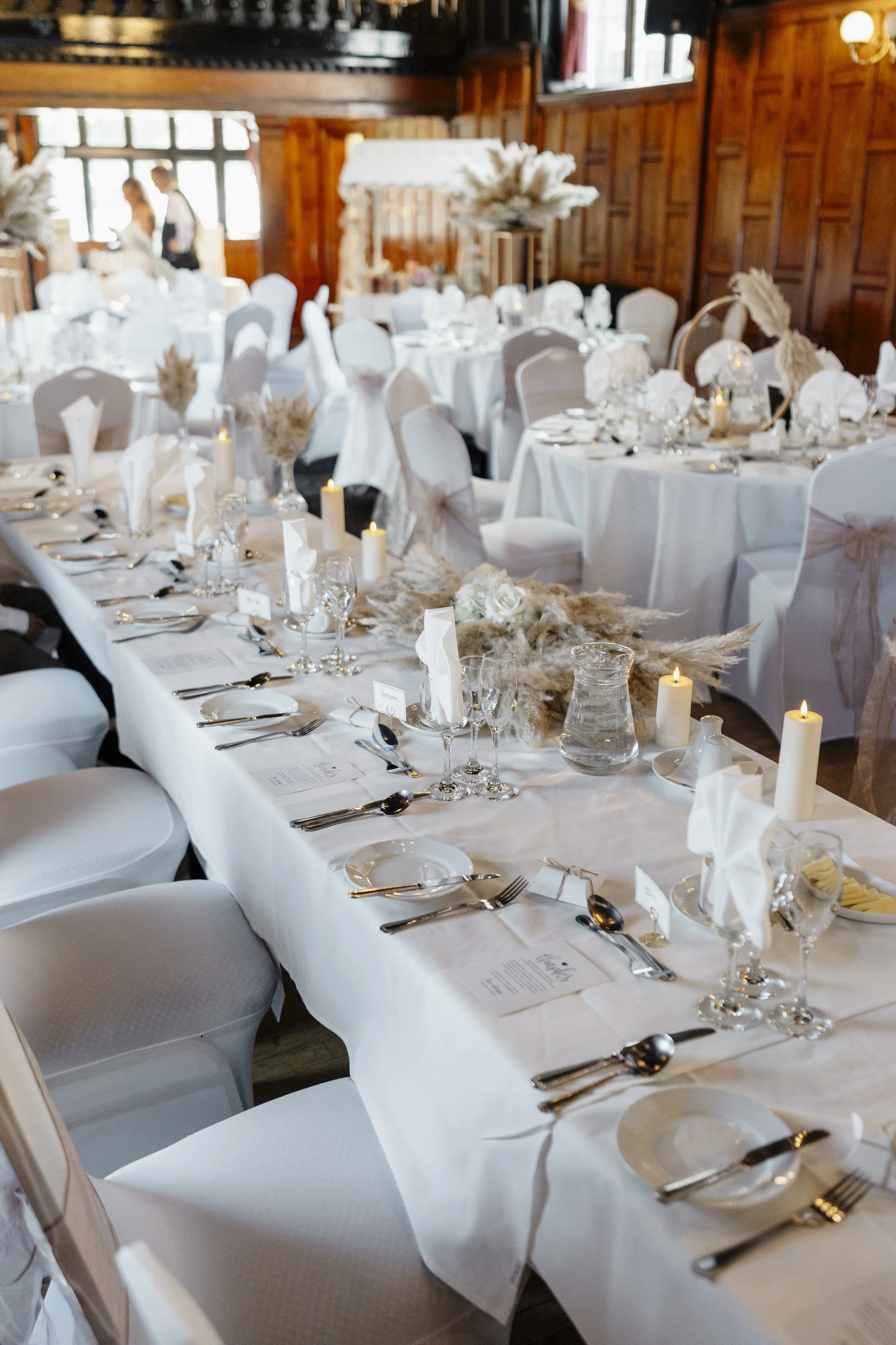 Elegant wedding reception with round tables covered in white tablecloths, decorated with candles, white floral arrangements, and place settings, in a wood-paneled room with large windows and couples in formal attire in the background.