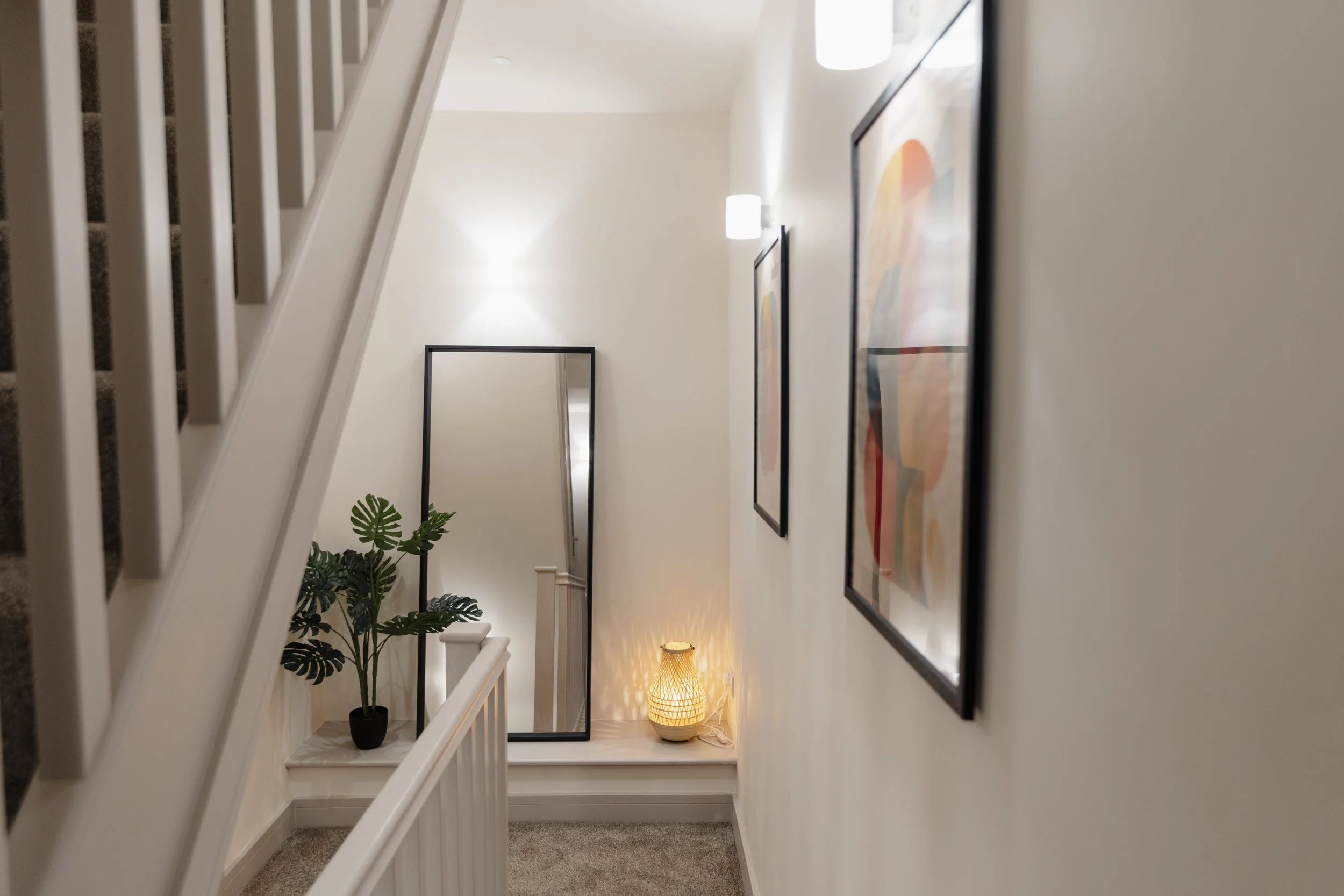 Hallway with framed artwork on the wall, a potted plant, a standing mirror, a lamp with a woven shade, and wall-mounted lights.