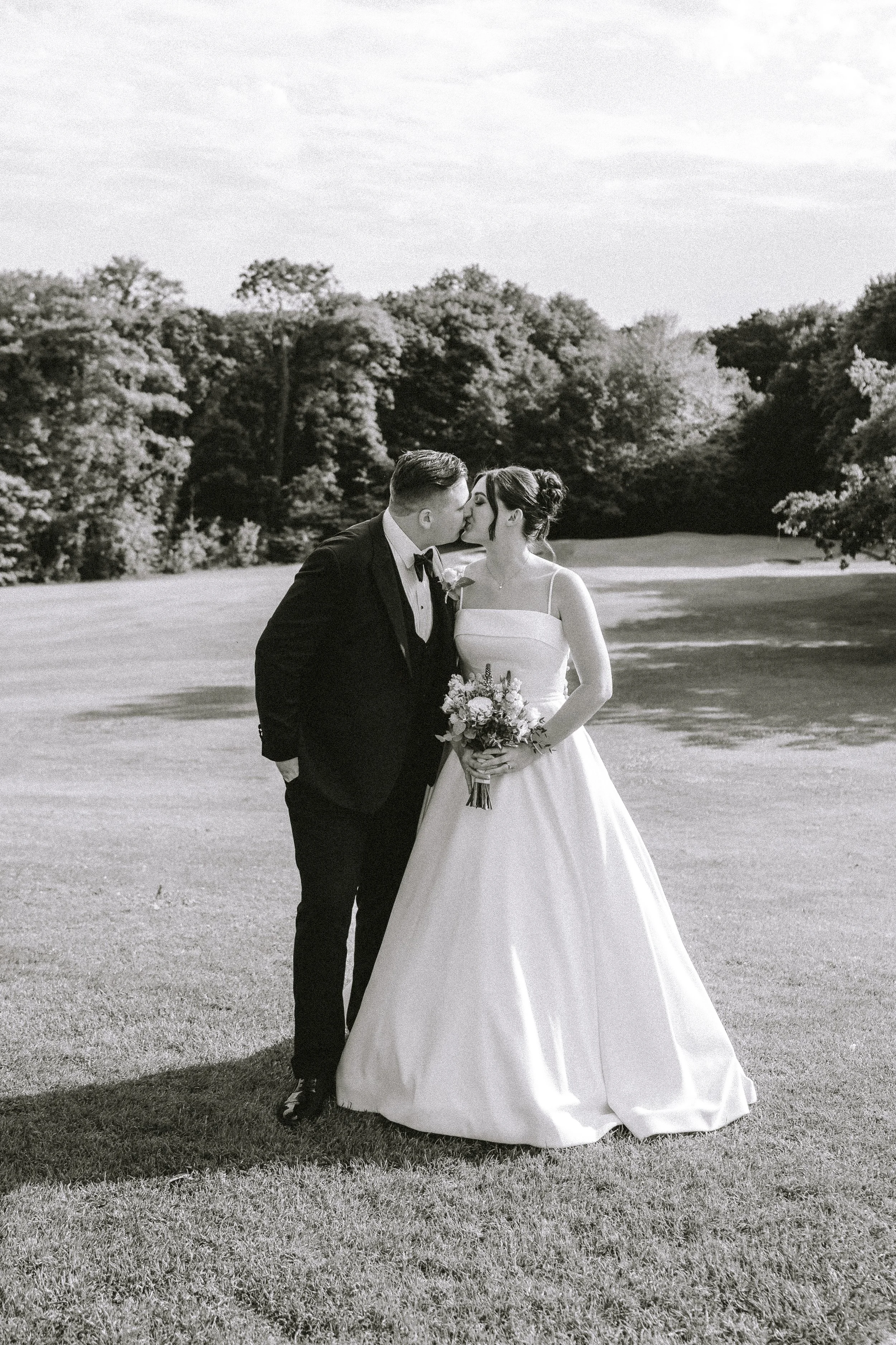Black and white photo of a bride and groom kissing outdoors, with trees and grass in the background.