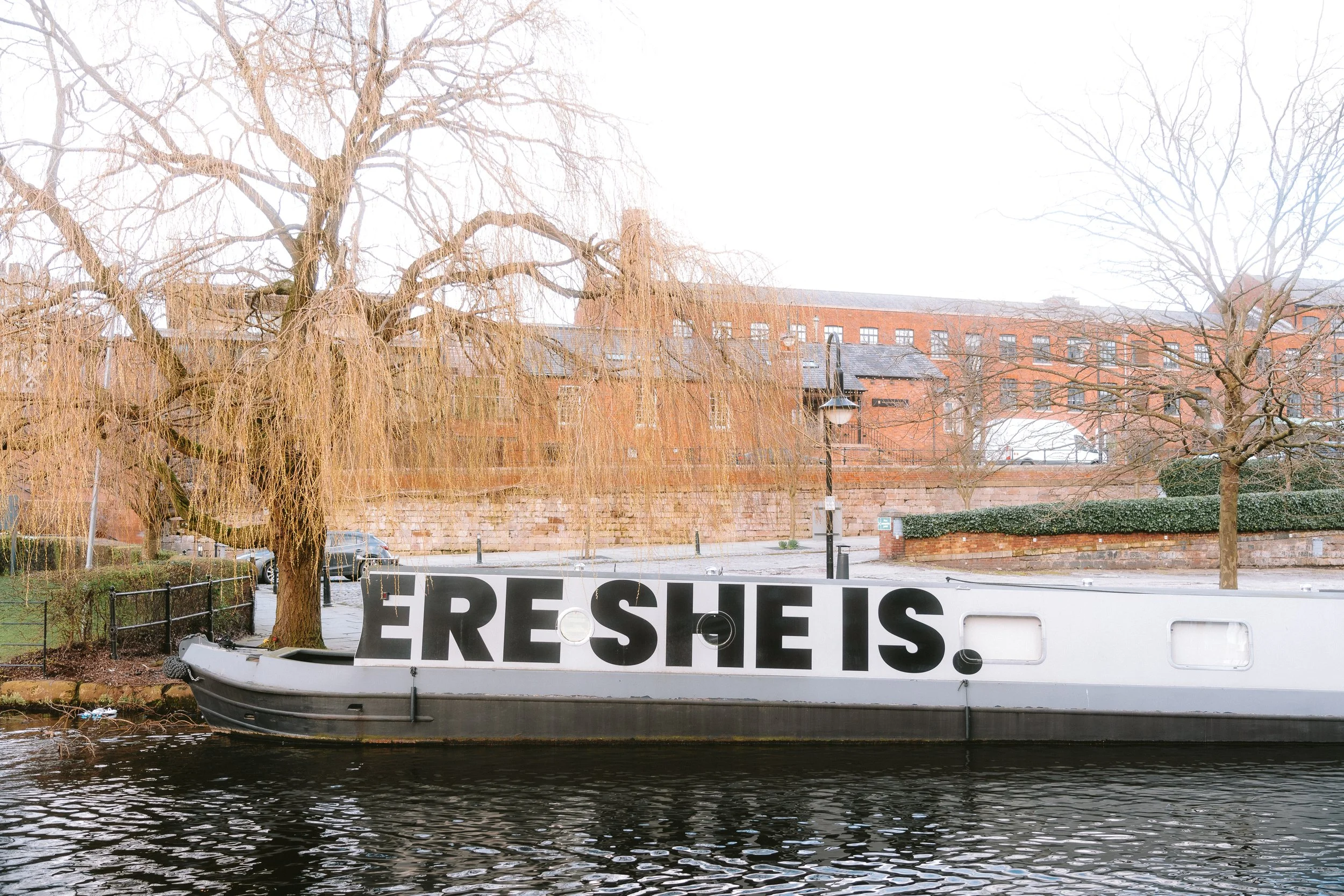 A boat with a large white sign that reads 'RESCUE IS' is docked on the water next to a riverside walkway. In the background, there's a large brick building, leafless trees, and a cloudy sky.