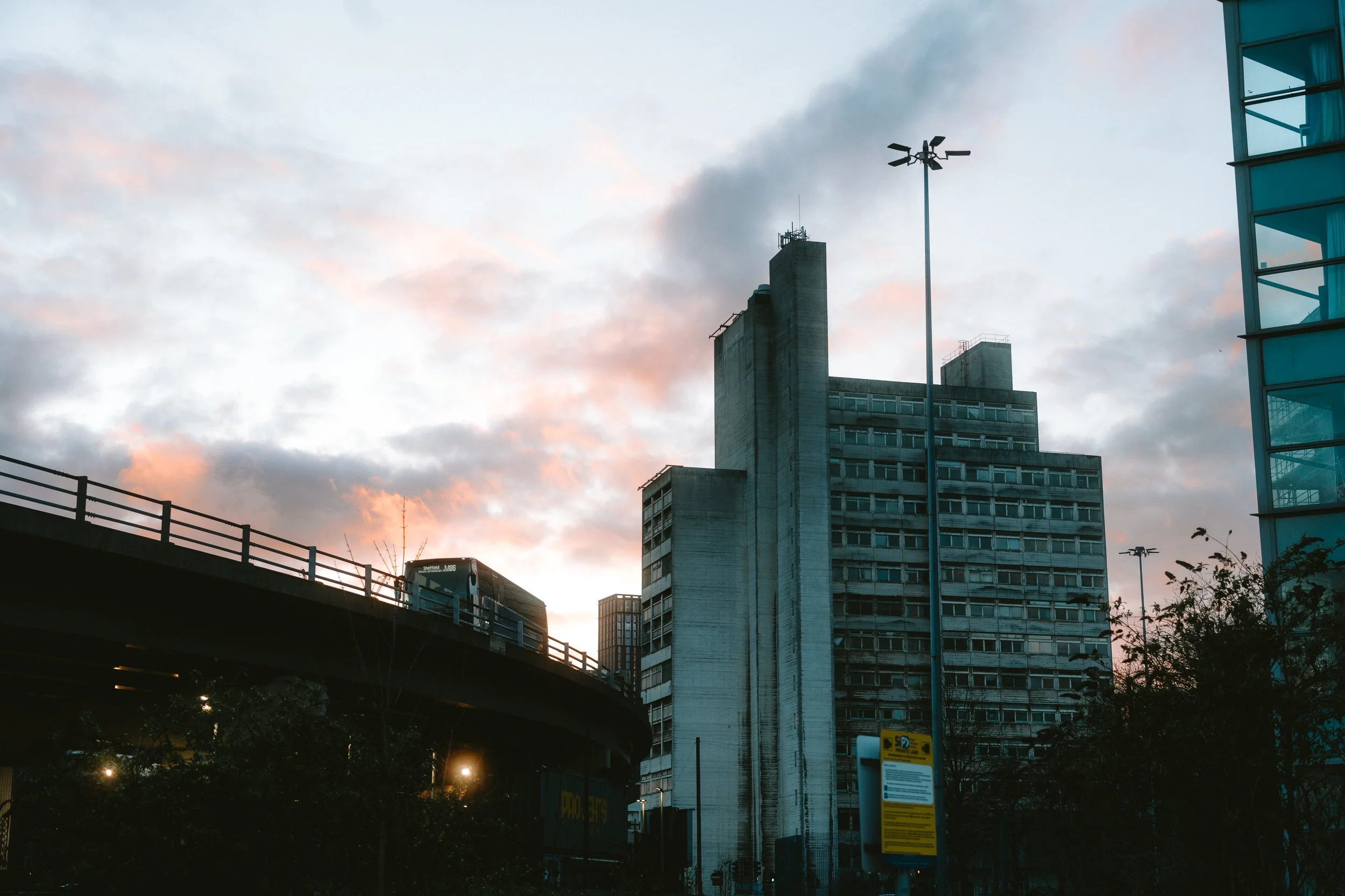 Cityscape at sunset featuring tall buildings, an overpass with a bus, modern glass structures, and a cloudy sky with pinkish hues.