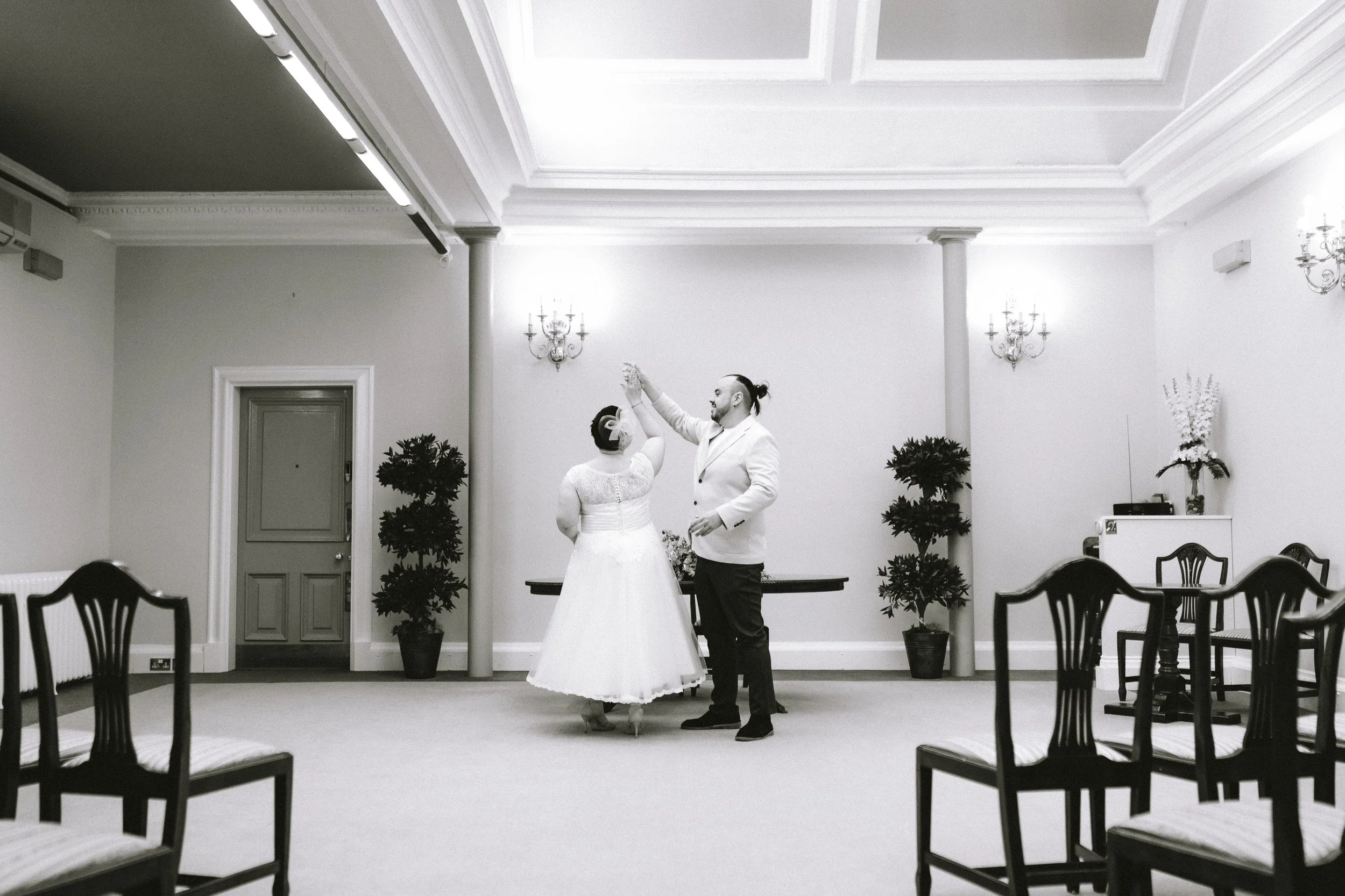 A bride and groom dancing in a decorated reception hall with empty chairs around them, potted plants, and chandeliers on the walls.