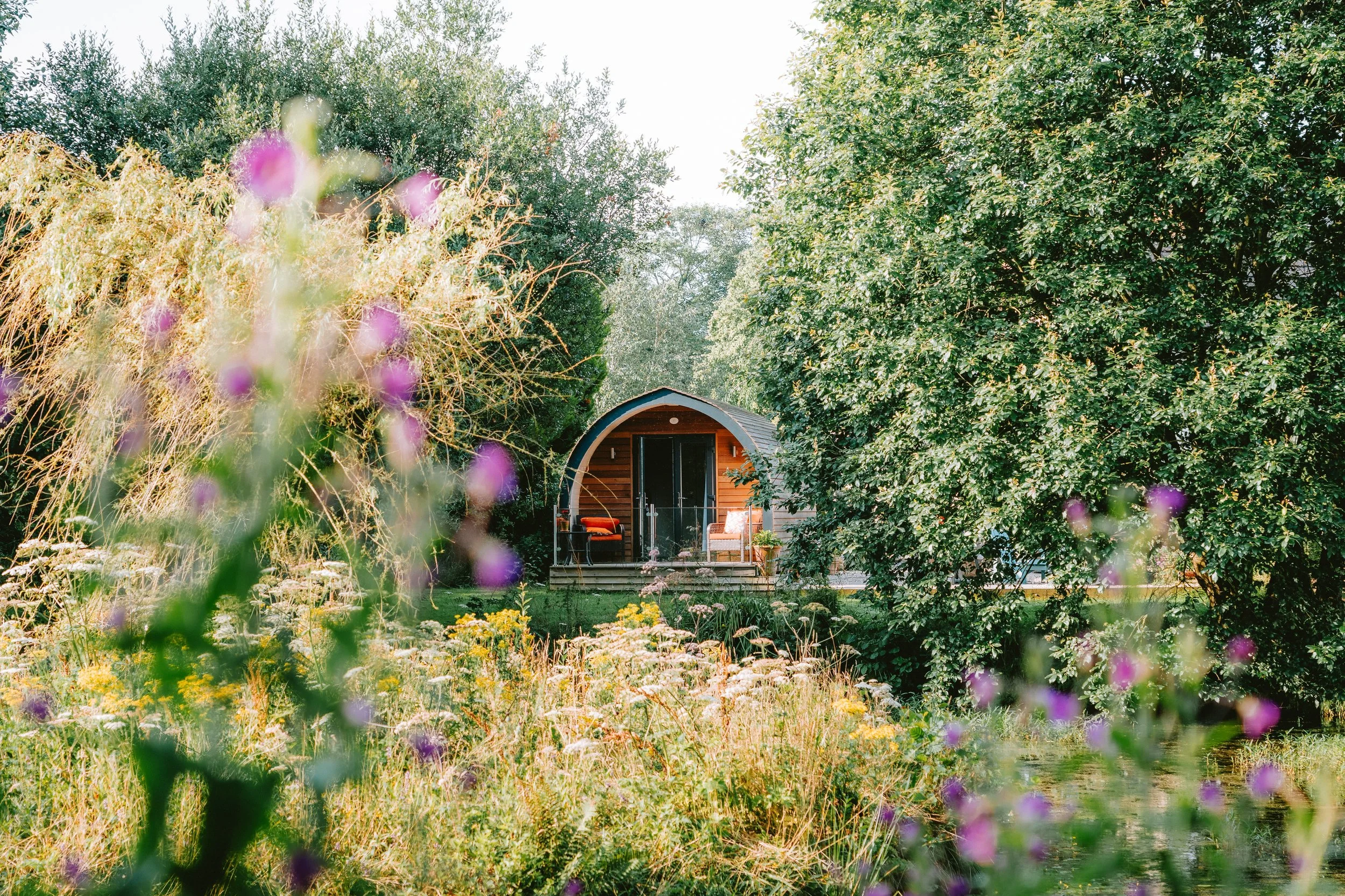 A cozy outdoor sitting area with chairs and a table on a small deck, surrounded by lush trees and colorful flowers in a garden.