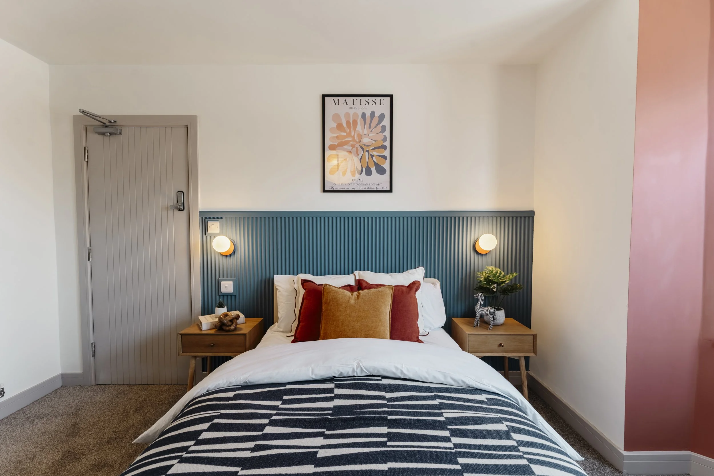 A bedroom with a bed featuring striped bedding, orange and beige pillows, wood nightstands on each side, one with a decorative rope sculpture, a small plant, and a zebra figurine. The headboard is painted blue with vertical grooves. Above the bed is 