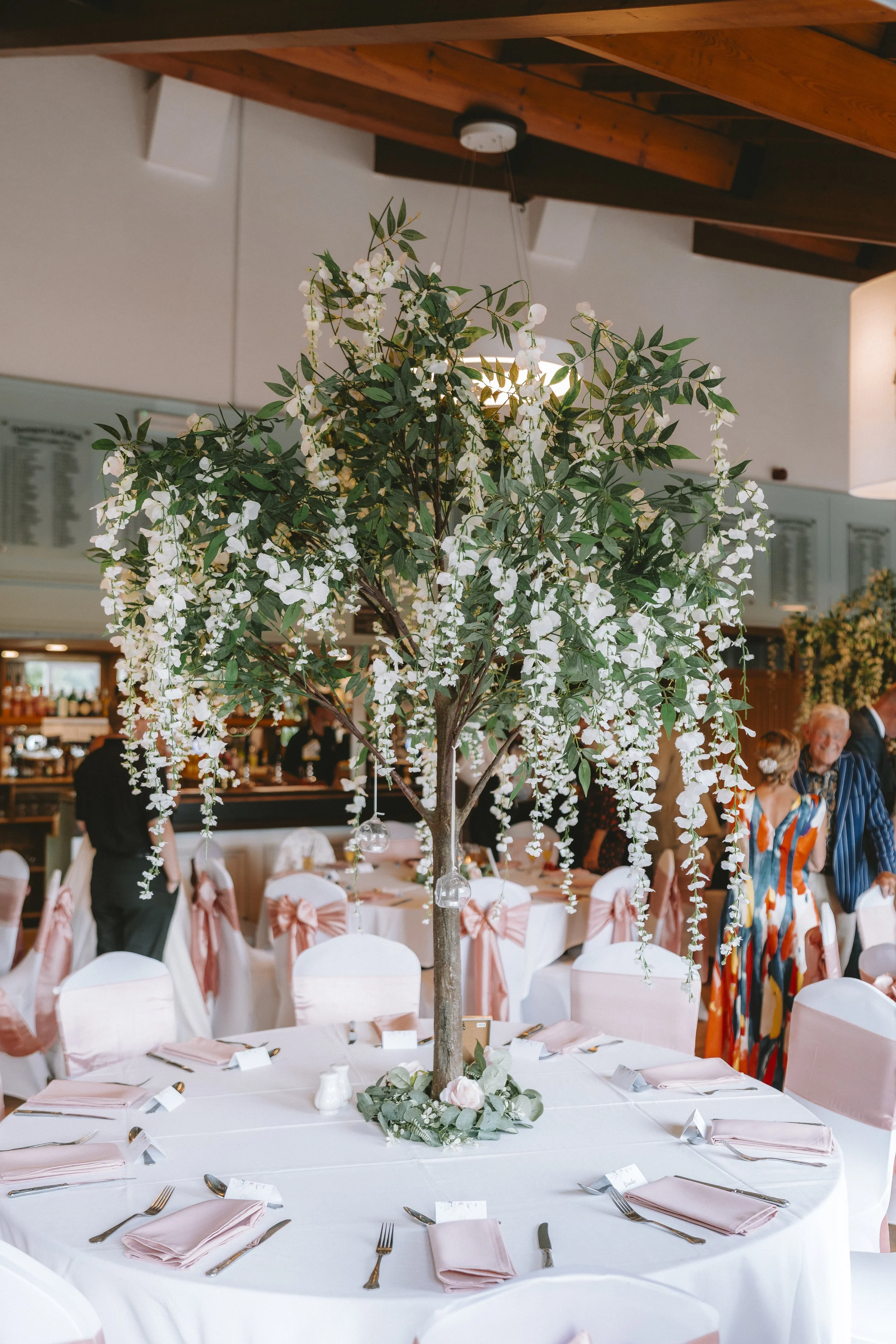 A large indoor celebration space with round tables covered in white tablecloths, set with silverware, pink napkins, and small name cards. The chairs are draped in white fabric with large blush pink bows. There is a tall decorative tree with white flo