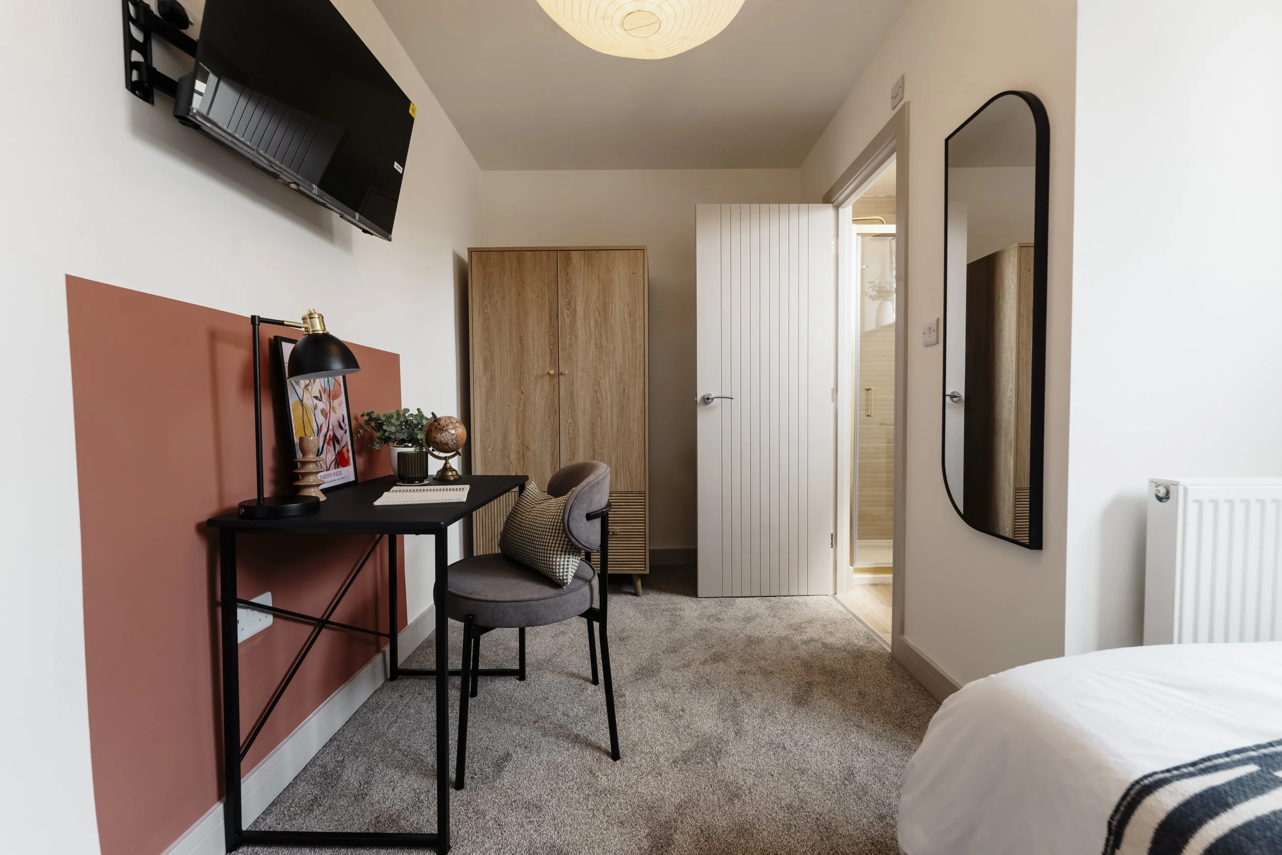 A bedroom with a black desk, a gray upholstered chair, a small plant, and framed artwork on a rust-colored accent wall. There is a wardrobe and a door leading to an ensuite bathroom. A wall-mounted TV and a tall mirror are also visible. The room has 