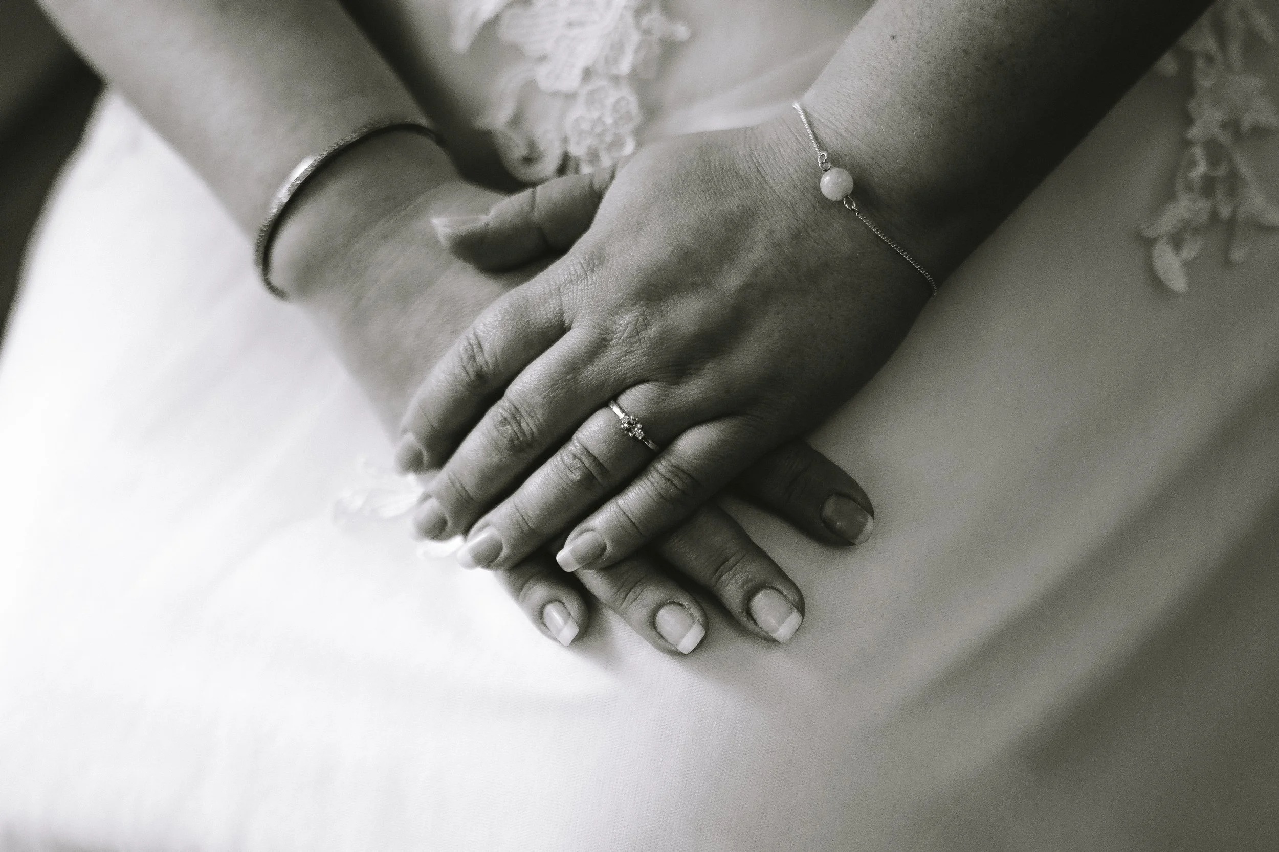 Close-up of two hands gently holding each other on a white surface. The person on the right is wearing a delicate bracelet with a white bead, and a ring on the ring finger. The person on the left is wearing a silver bracelet.