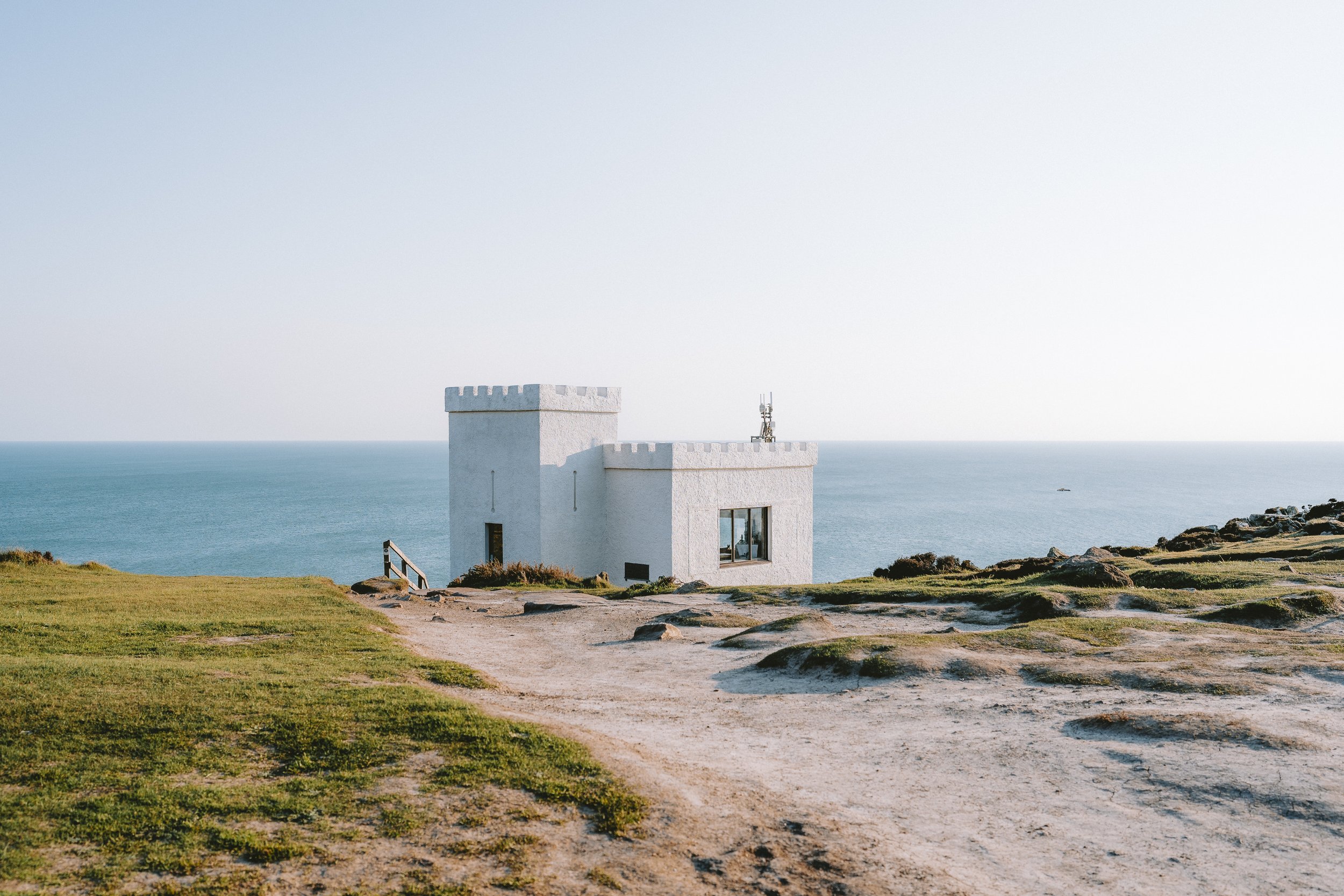 A white castle-like building with battlements on a grassy hill overlooking the ocean, with a clear blue sky in the background.