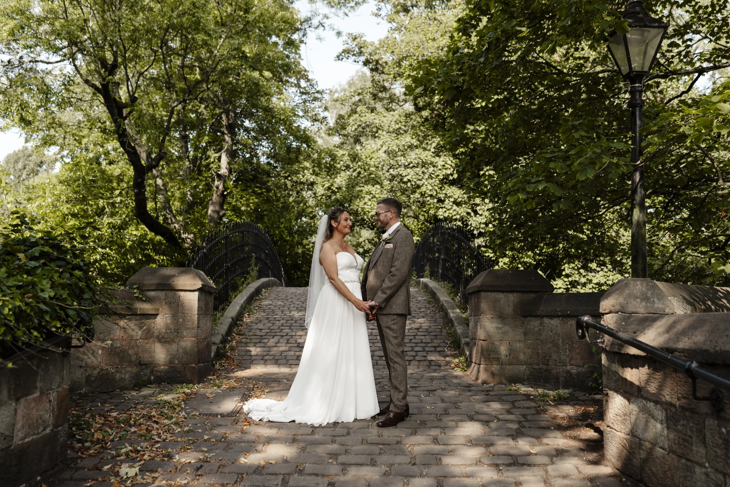 A bride and groom standing hand in hand outdoors on a cobblestone bridge, surrounded by green trees and a black iron fence, with a lamppost nearby.