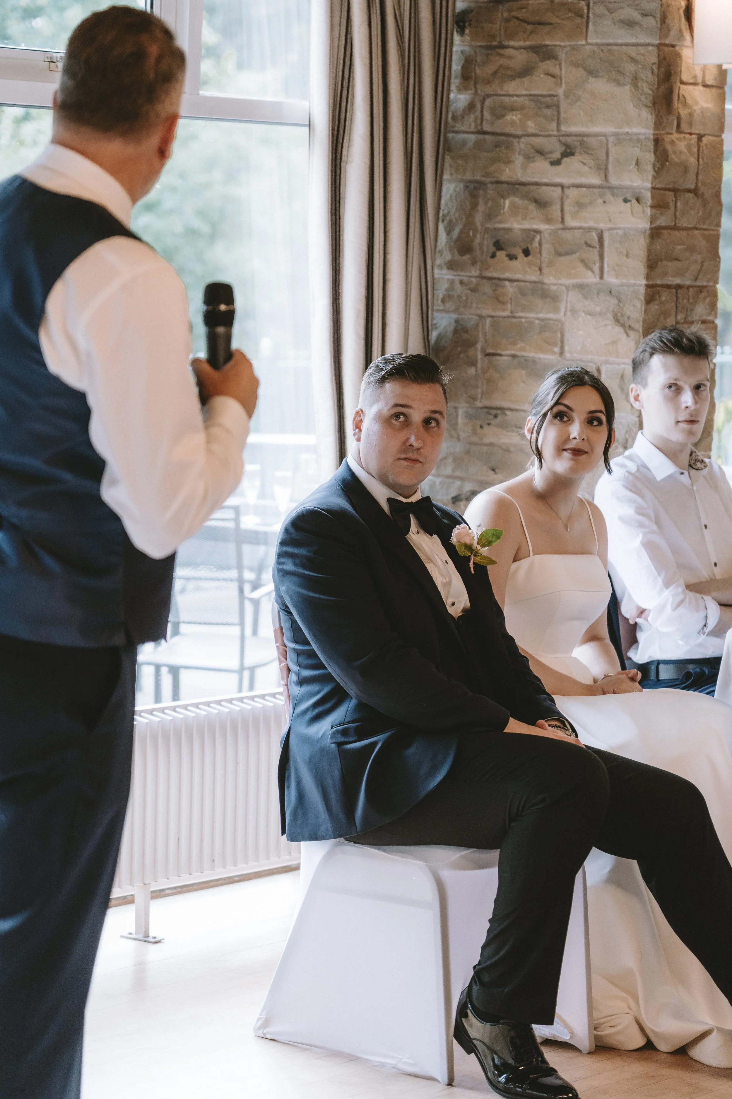 Man giving a speech at a wedding reception with three seated guests listening attentively.