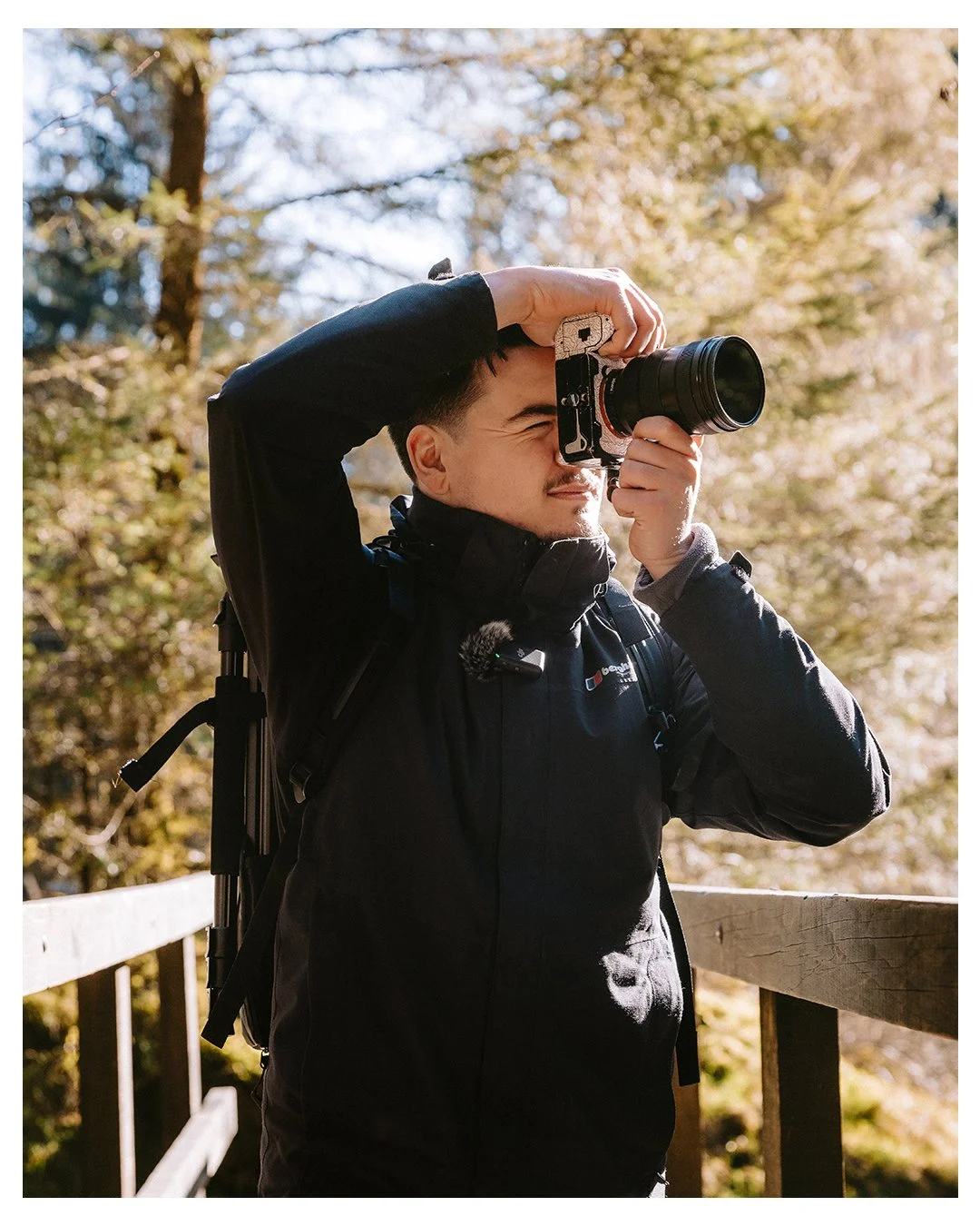 A man standing outdoors on a wooden bridge taking a photo with a camera, surrounded by trees with autumn-colored leaves.