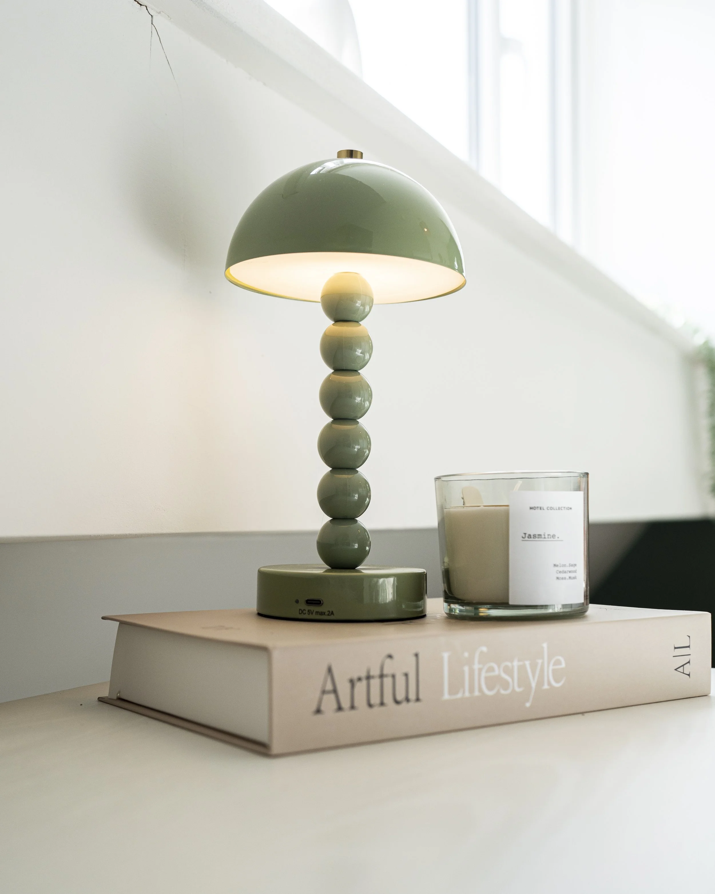A green lamp with a dome-shaped shade and a base made of stacked rounded shapes, placed on a light-colored surface. To the right of the lamp, there is a transparent candle holder with a white candle inside, a label reading "Jasmine," and partial text