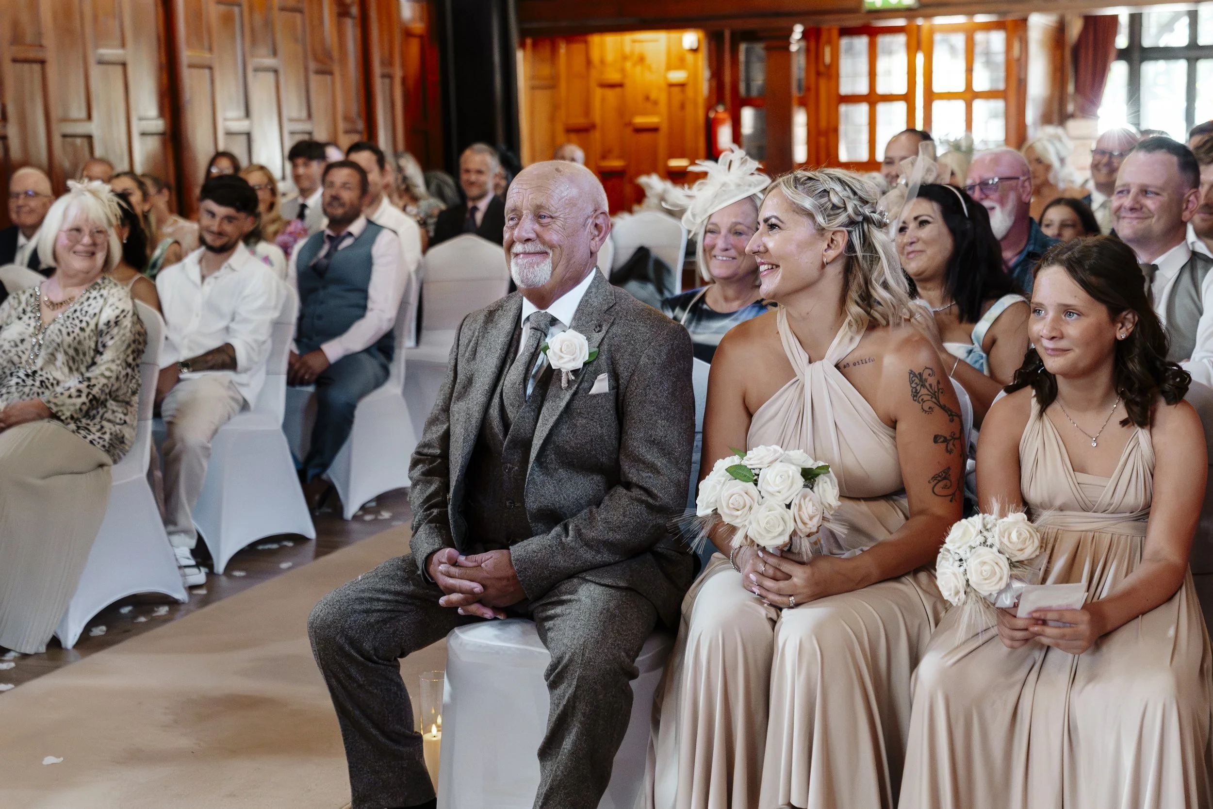 People dressed in formal attire sitting in a wooden-paneled wedding ceremony hall, smiling and listening to the ceremony.