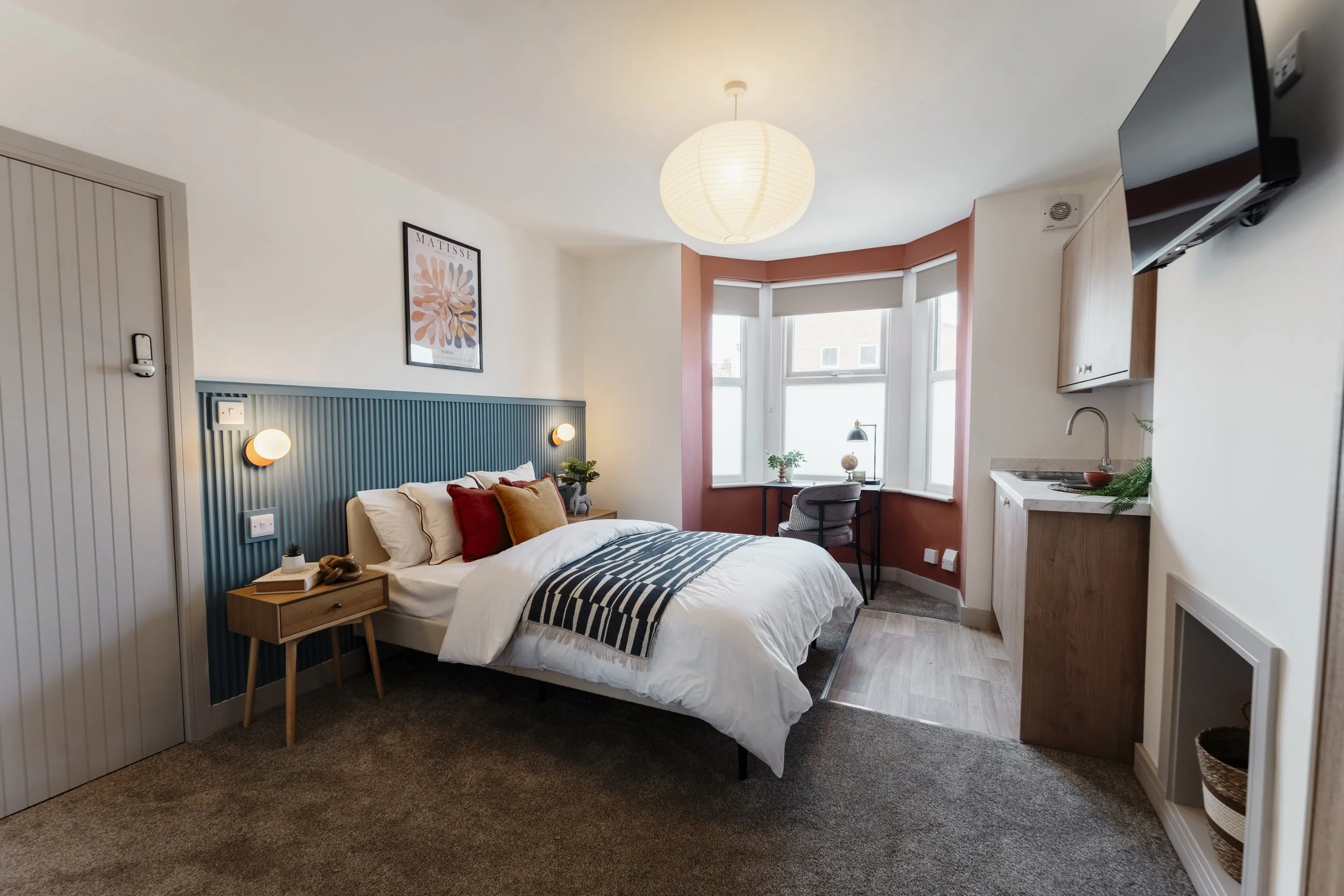 A cozy bedroom with a large bed, colorful pillows, bedside table, and wall-mounted reading lights. The room has a corner window with a desk and chair, a lamp, and plants. A paper lantern ceiling light hangs above.