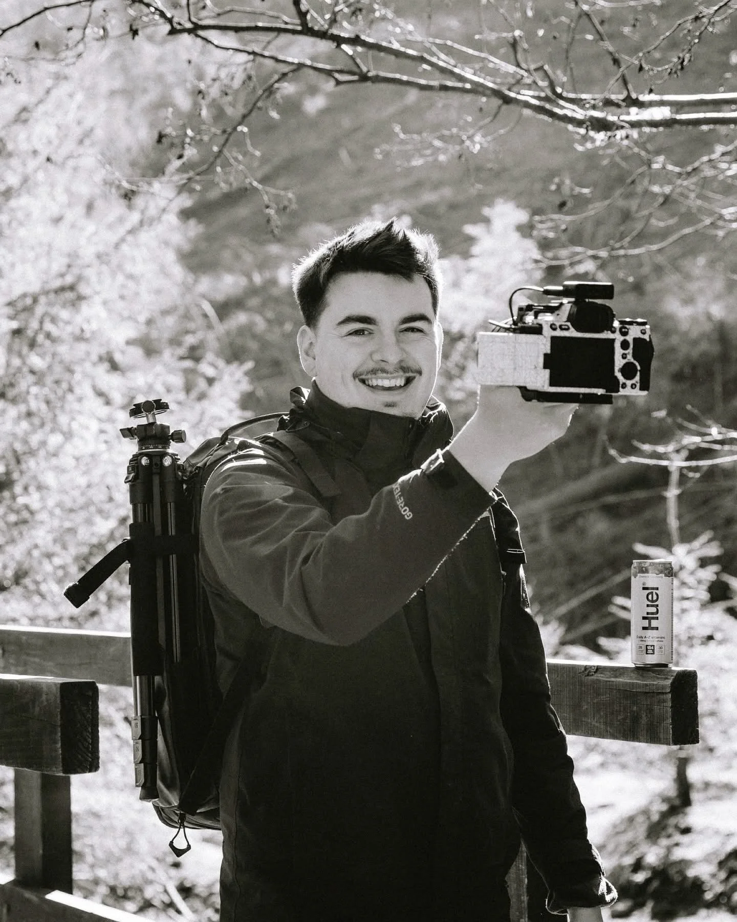 Glynn Hodsonn with short dark hair and a goatee holding a camera outdoors, with a backpack on his back and a can of Heiß energy drink placed on a wooden railing next to him, surrounded by trees and sunlight.