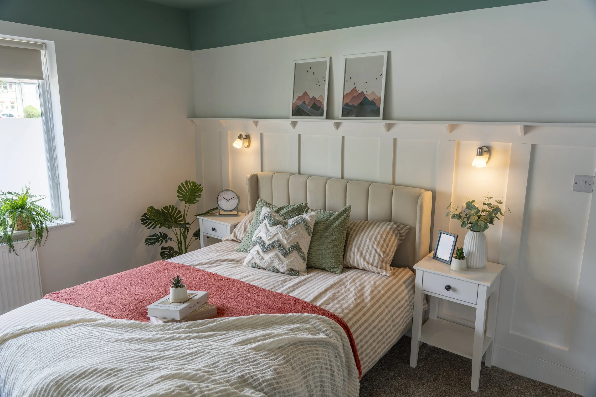 A bedroom with a bed decorated with pillows and a throw blanket, two white nightstands with decor, plants, and a clock, framed mountain print art above the bed, wall-mounted lamps, and a window with a white blind, in a modern, cozy style.