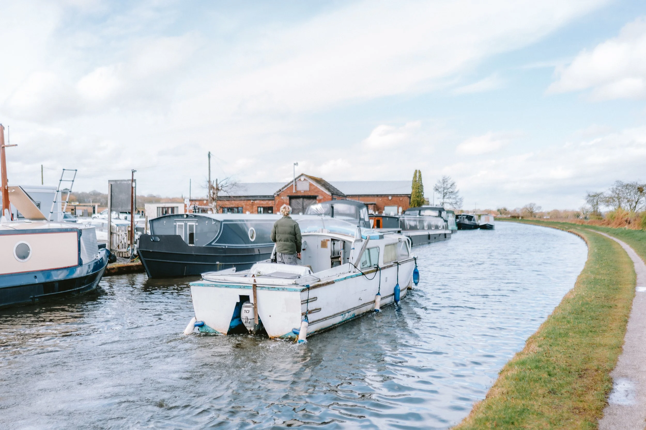 Man walking along a canal with boats moored on the water and a brick building in the background.