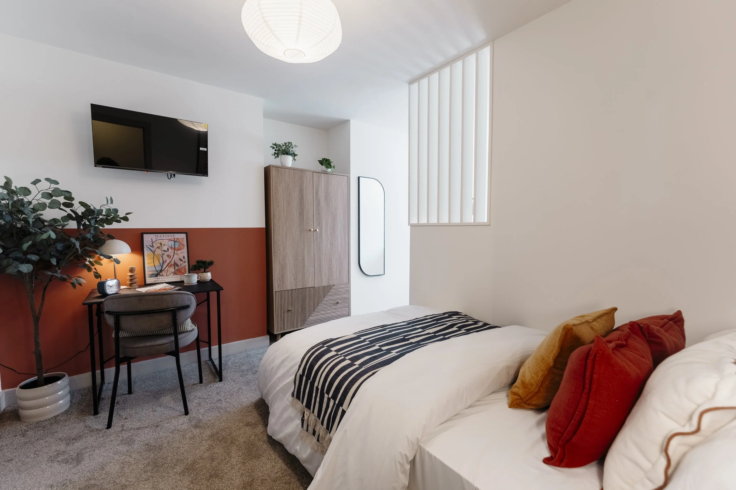 A bedroom with a bed that has white bedding and orange pillows, a dresser with plants on top, a mirror on the wall, and a small desk with a chair, picture frame, and various decorative items, illuminated by ceiling and table lamps.