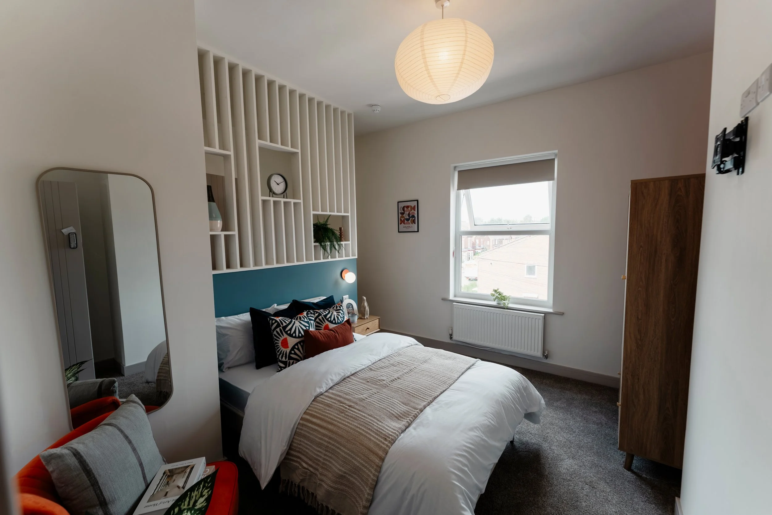A bedroom with a bed, decorative pillows, a wooden wardrobe, a window with a blind, wall decorations including a clock and a framed art piece, a paper lantern ceiling light, a mirror, and a small table with a plant and some items.