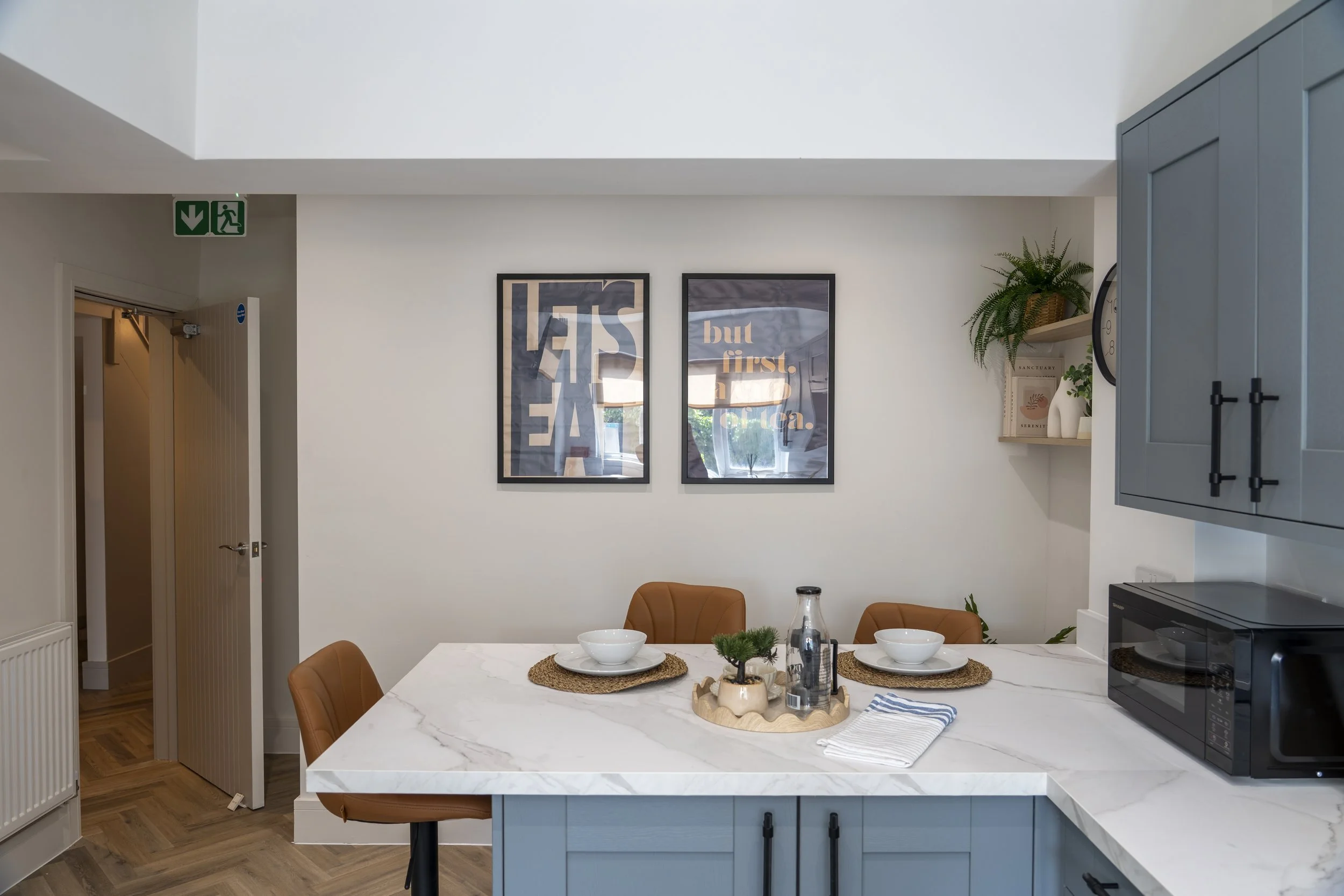 Interior of a modern kitchen and dining area with a white marble countertop island, blue cabinets, framed wall art, plants, and a microwave.