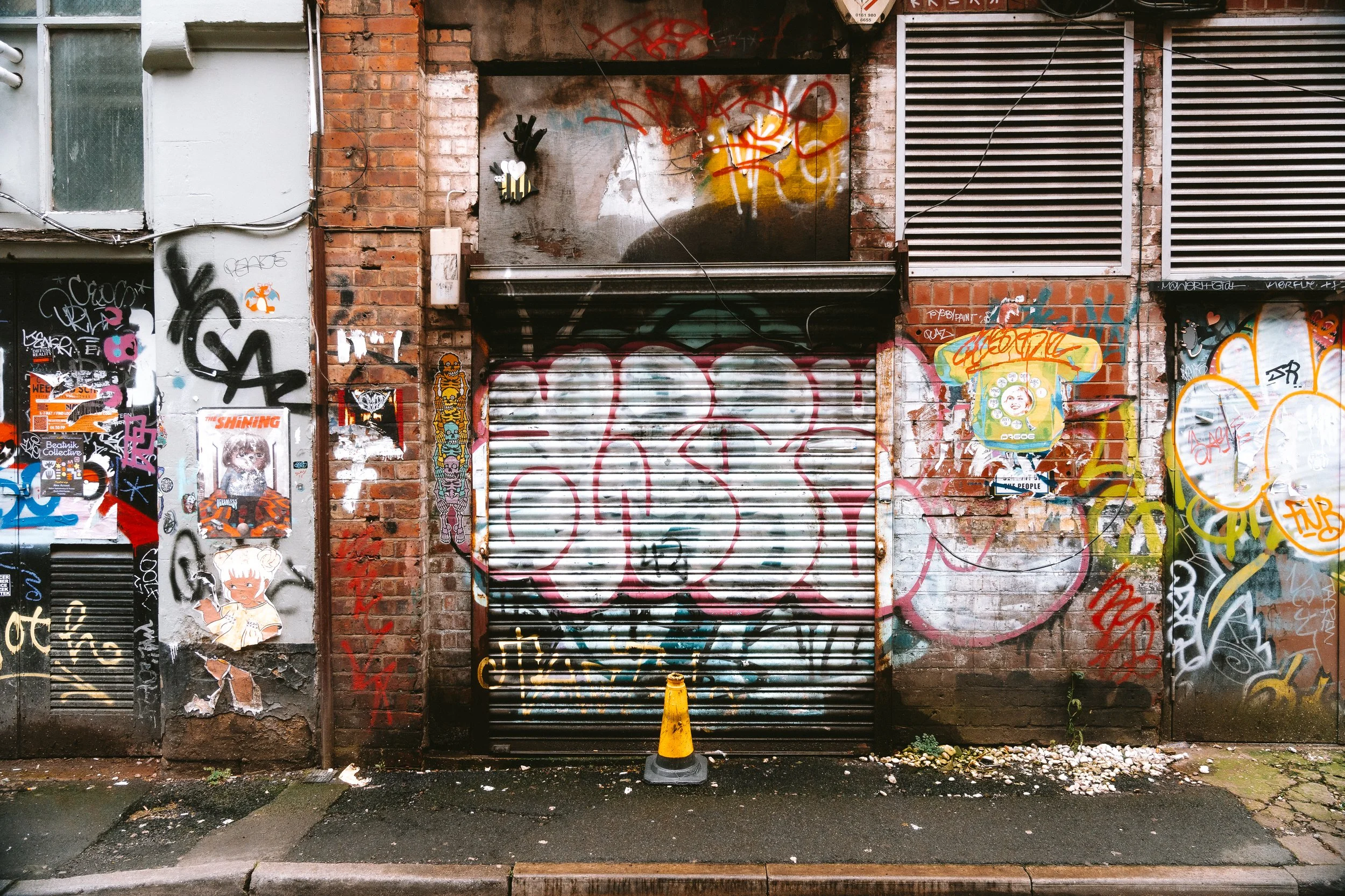 A brick building facade covered with colorful graffiti, street art, and posters. There is a metal roll-up door with a pink spray-painted design, a small decorative black hand with a white heart, and a yellow traffic cone in front of the door. The are