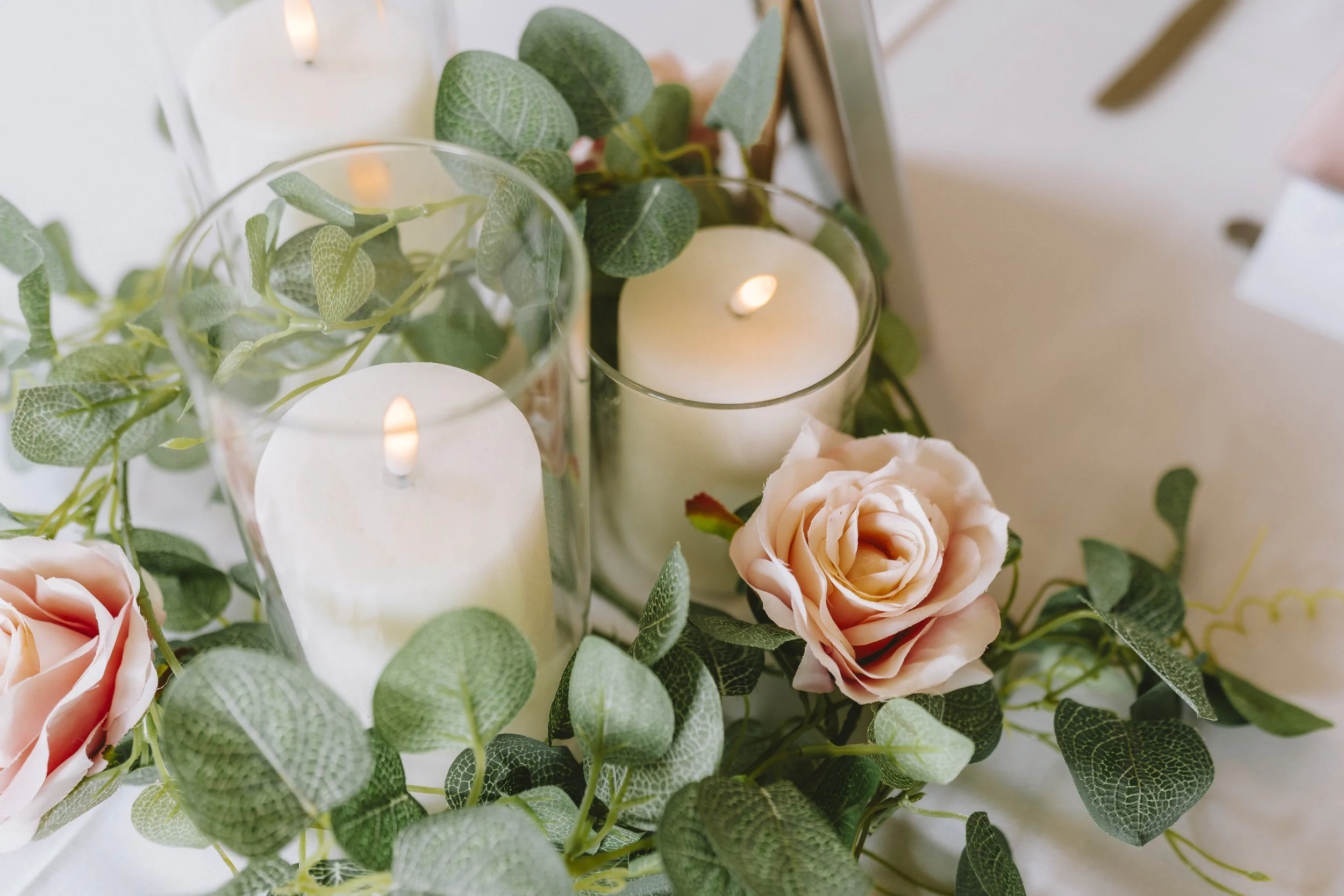 The image features a decorative arrangement of pale pink roses, green foliage with textured leaves, and white candles in glass holders, creating a soft and elegant display.