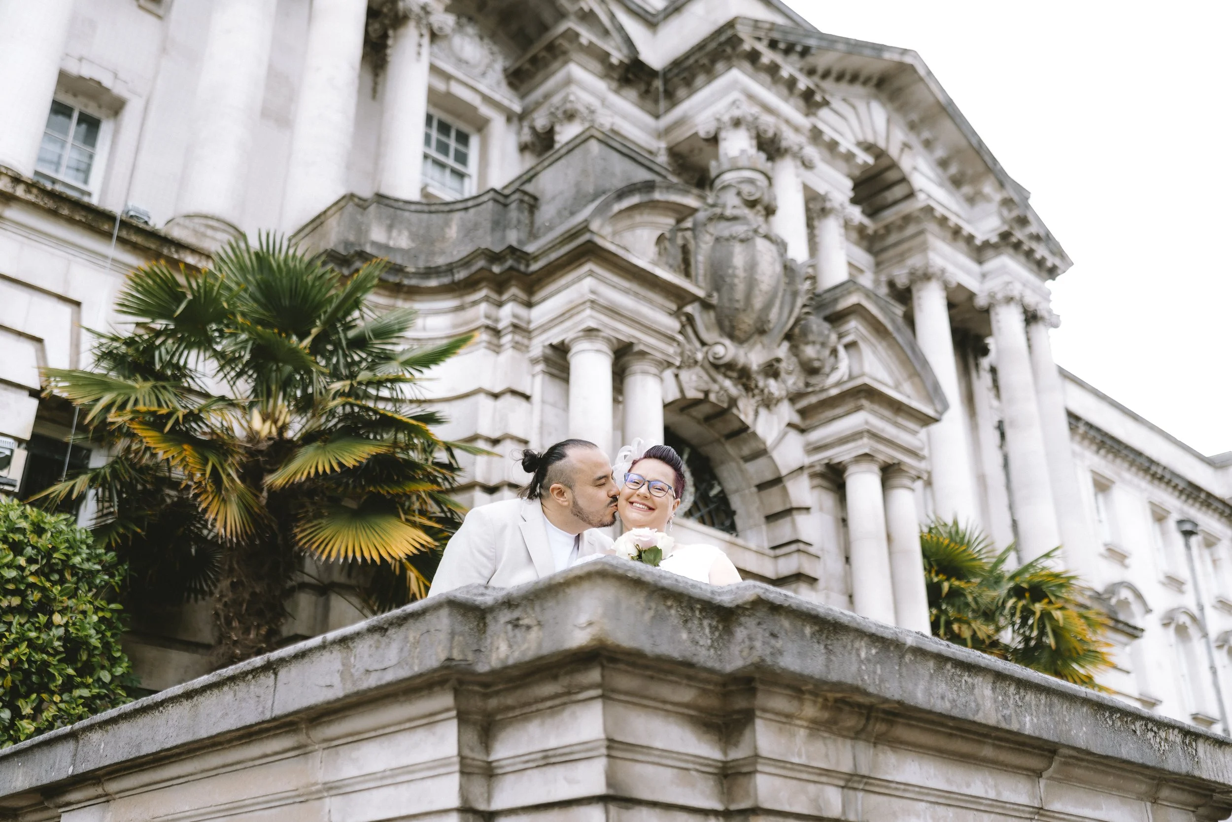 A couple in wedding attire sharing a kiss on a stone balcony in front of a historic stone building with ornate architectural details and columns, surrounded by greenery.