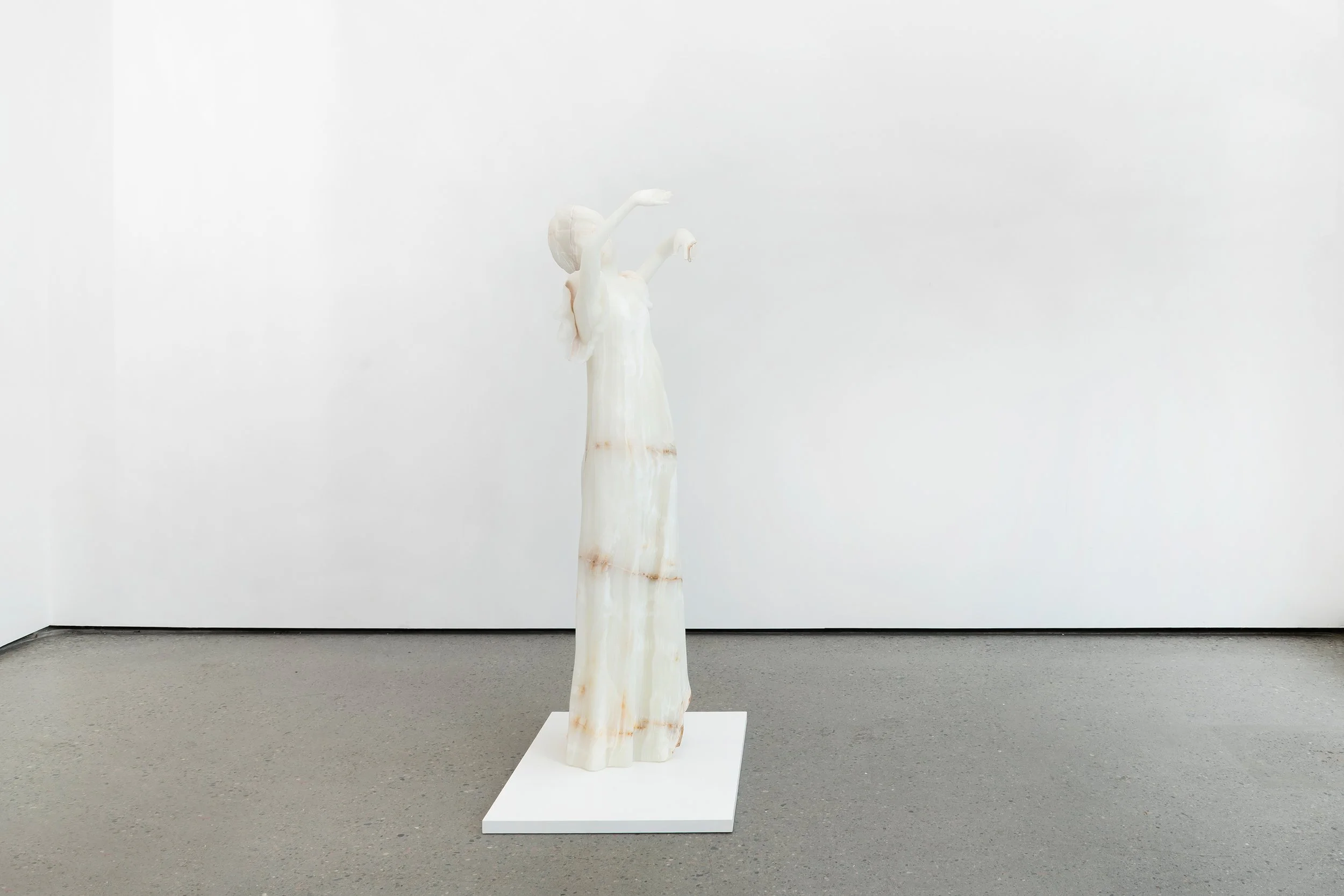 A sculpture of a woman with elongated arms and neck, standing on a white pedestal in a minimalist gallery space with white walls.
