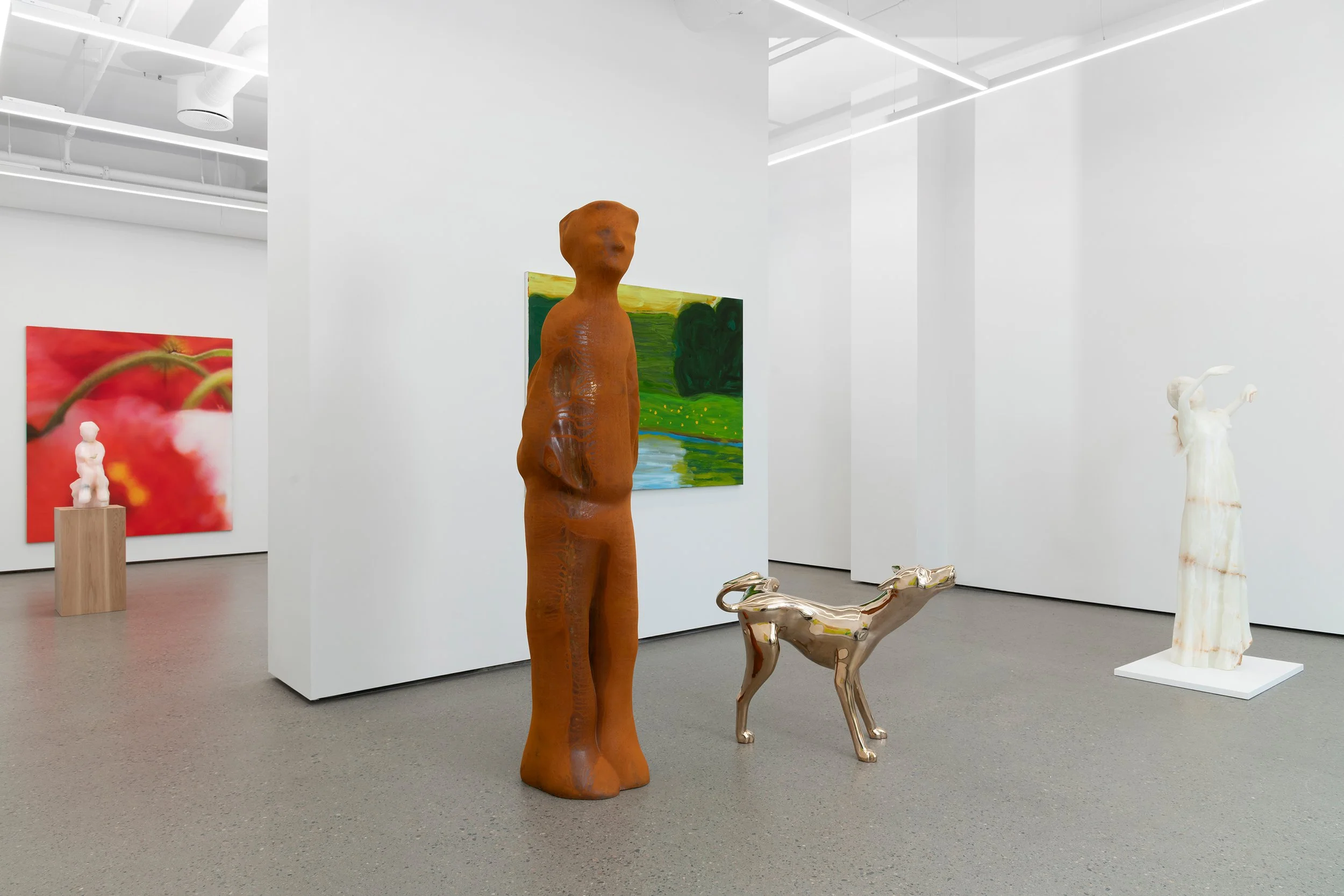Modern art gallery with sculptures and paintings. A tall, brown, human-like sculpture with a rounded head stands next to a shiny metallic dog sculpture. A white abstract sculpture with pointed arms is on a white platform. Two paintings, one with a re