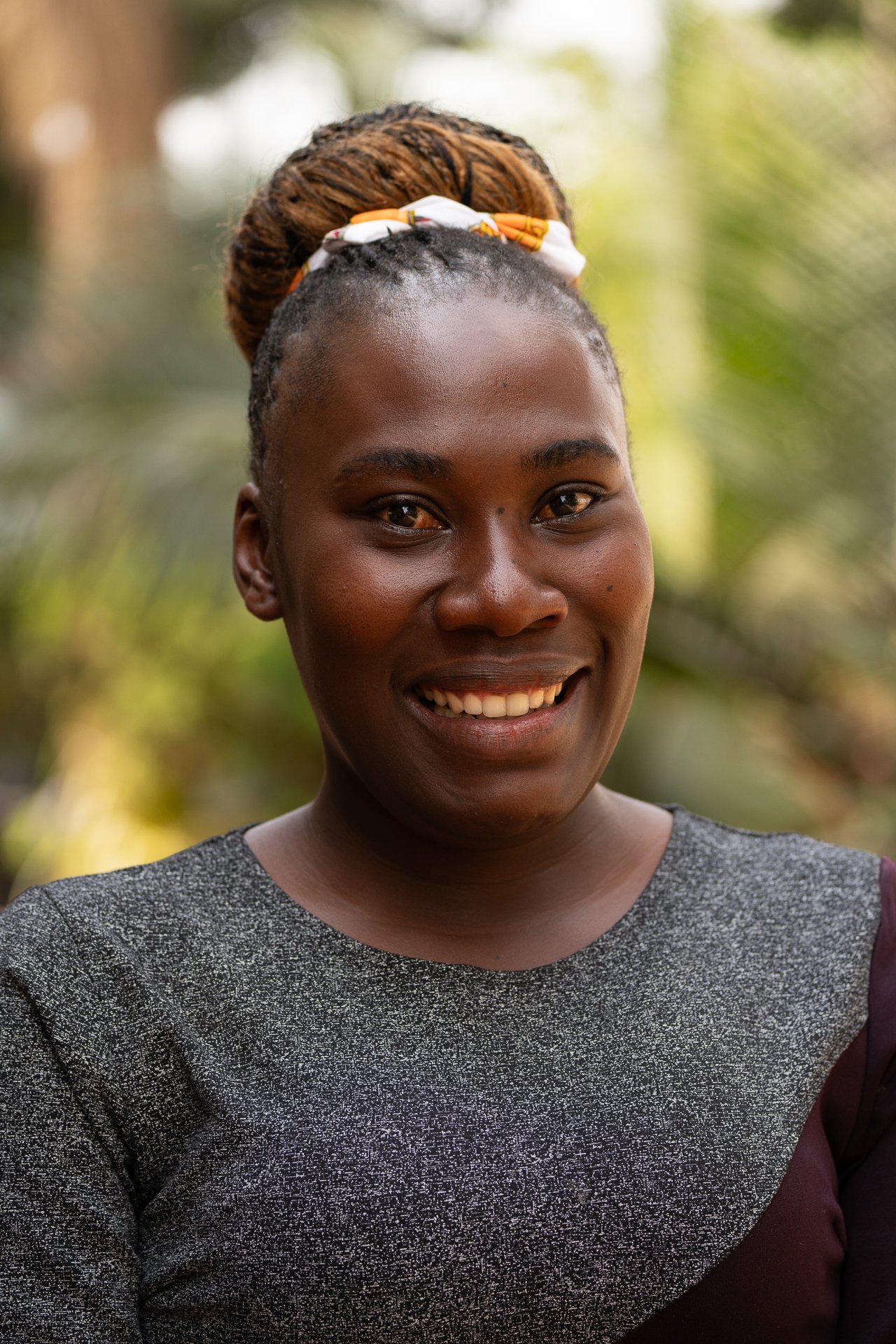 Lächelnde afrikanische Frau mit geflochtenen Haaren, die mit Band zusammengehalten sind, im Freien mit grüner Natur im Hintergrund.