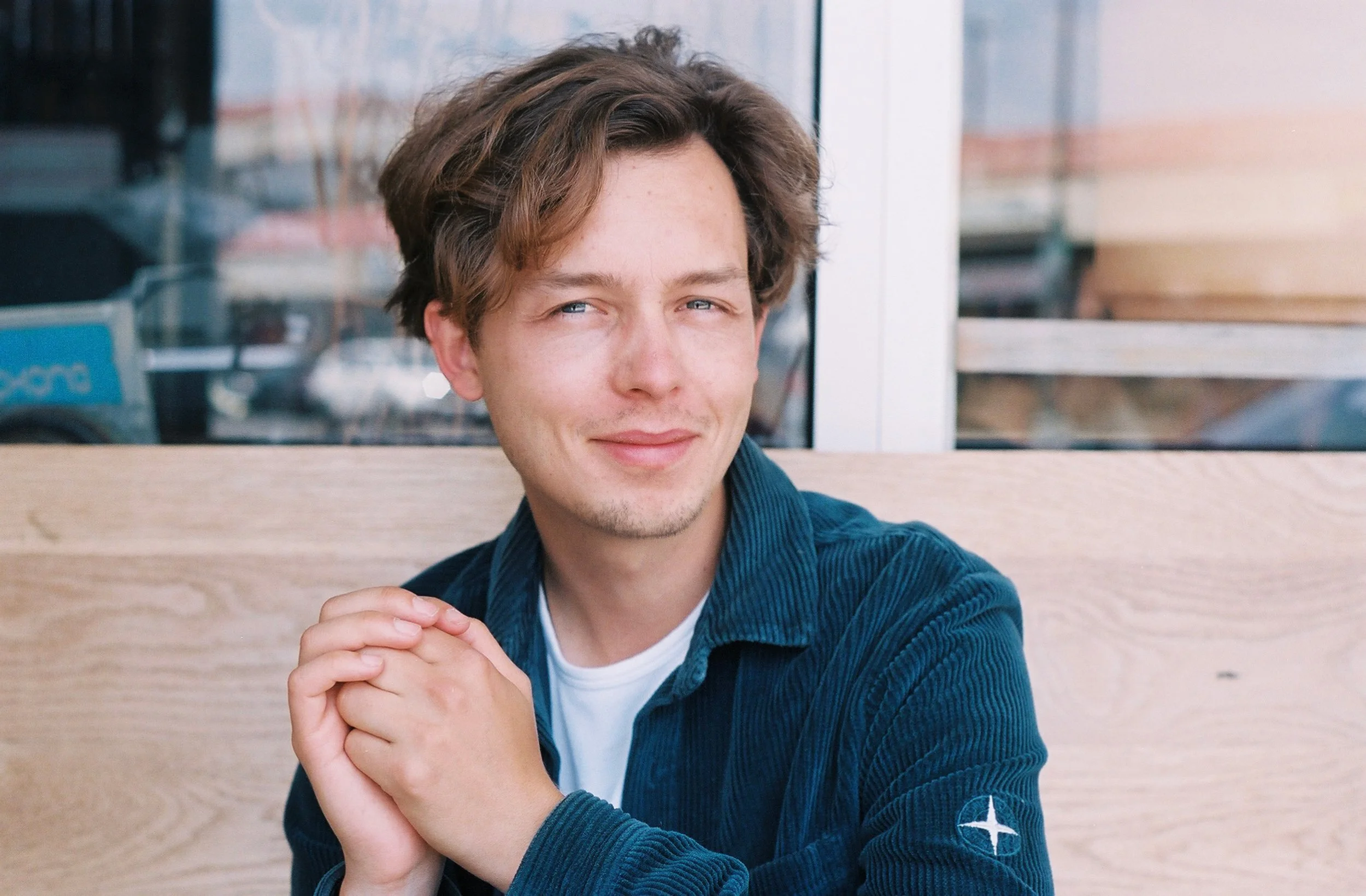 Junge Mann mit braunen, lockigen Haaren, blauen Augen, im Café sitzend, trägt eine dunkelblaue Jacke mit Logo auf dem Arm und hält die Hände zusammen.