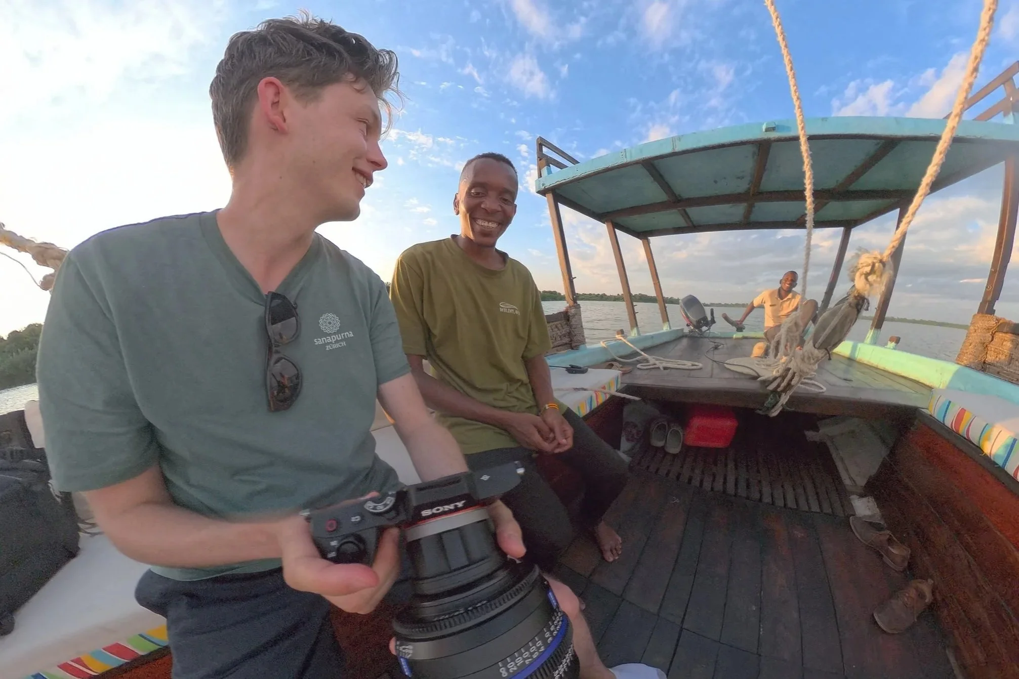 Drei Männer lachen und schauen sich auf einem Boot an, während das Wasser und der Himmel im Hintergrund zu sehen sind.