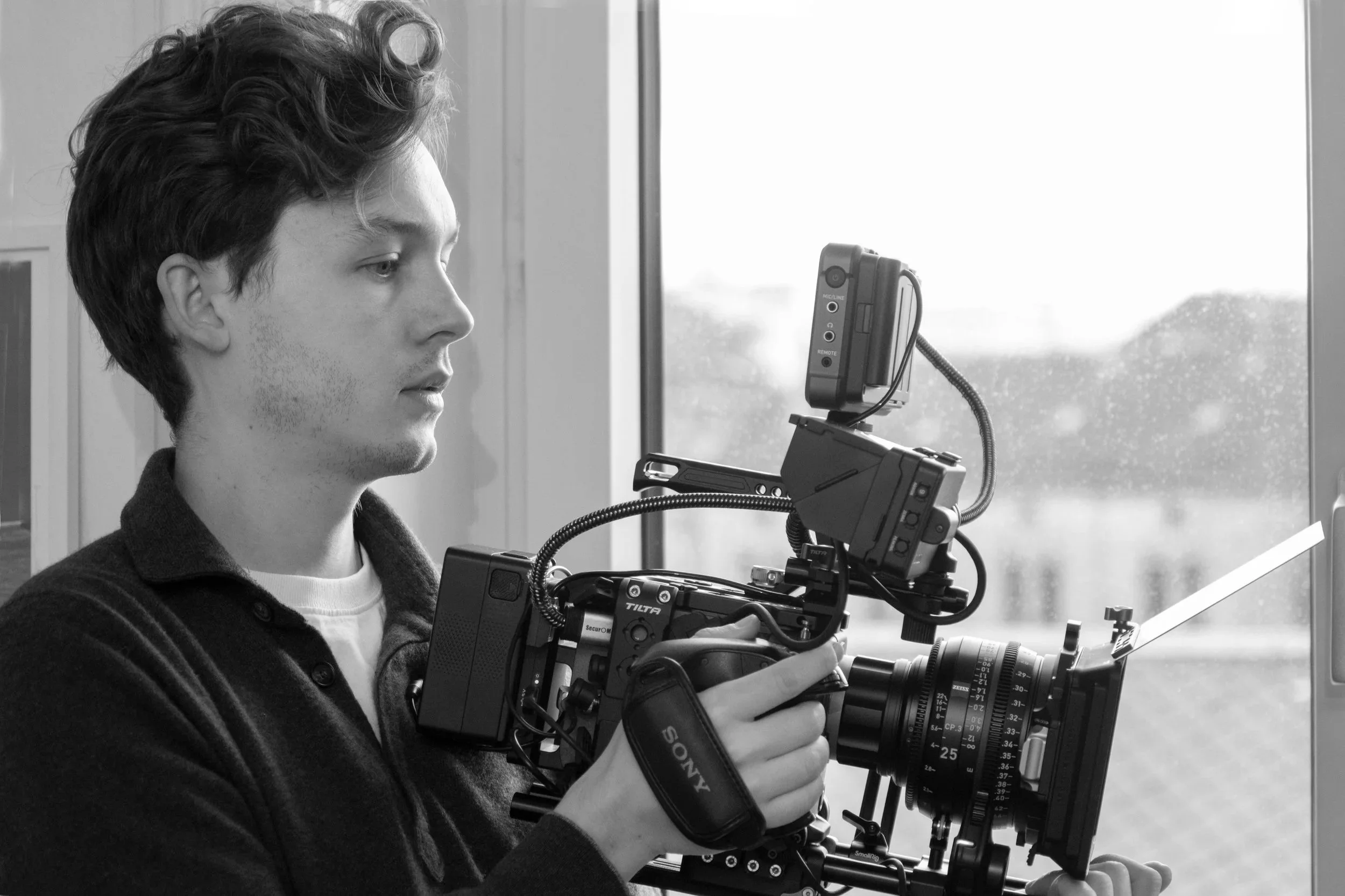 Ein junger Mann mit dunklen, lockigen Haaren, der mit einer Kamera auf einem Stativ vor einem Fenster steht und an der Kamera arbeitet.