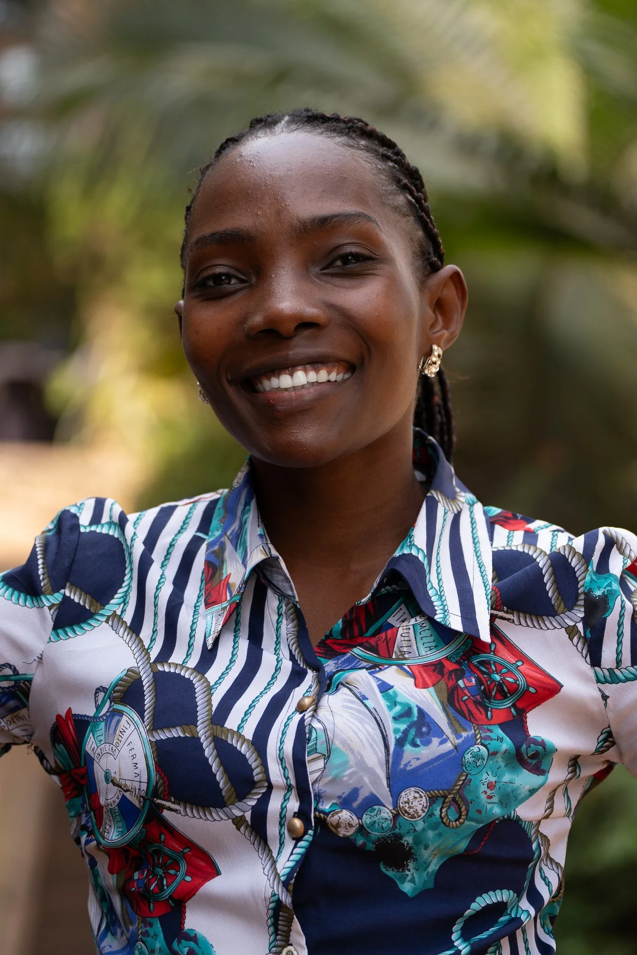 Lächelnde afrikanische Frau mit Zöpfen und Ohrringen, trägt ein buntes, gemustertes Hemd, im Freien vor grüner Hintergrundvegetation.
