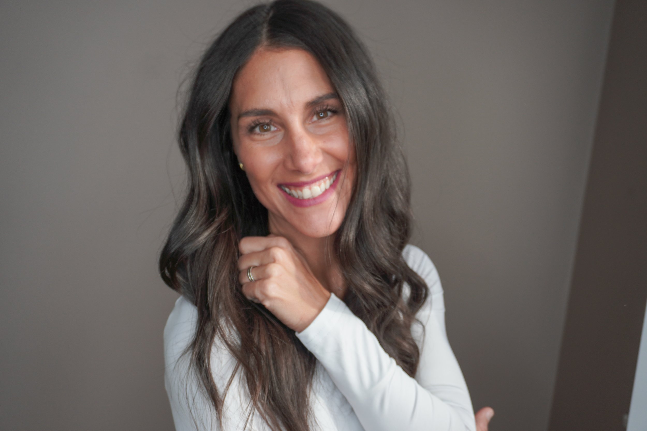 A woman with long, wavy dark hair smiling and looking at the camera, wearing a white long-sleeve top and standing against a plain background.