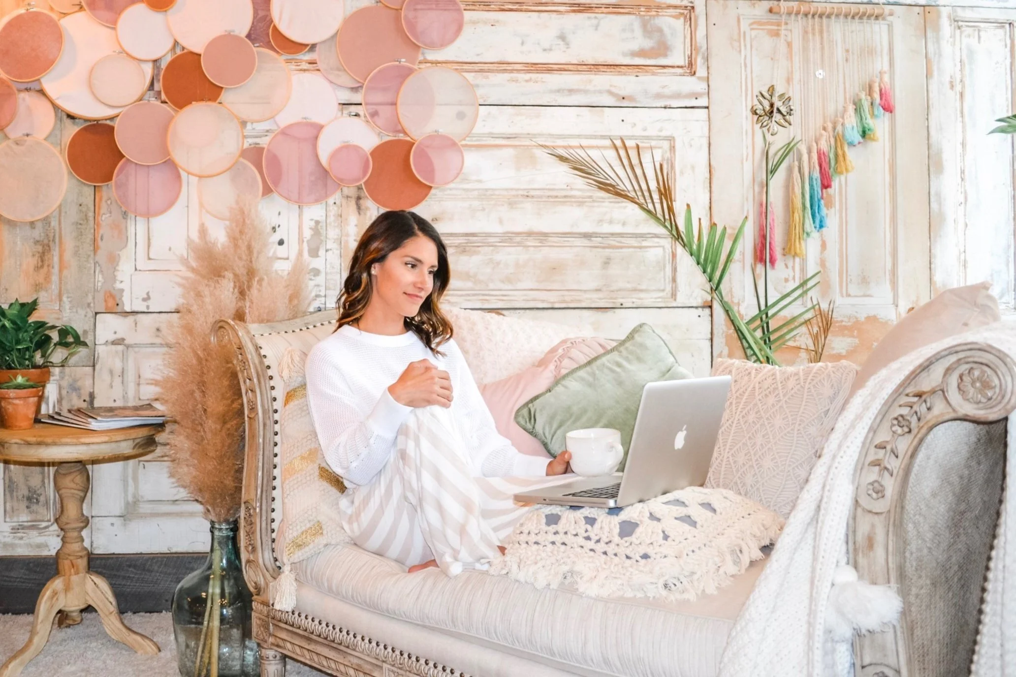 A woman sitting on a vintage beige sofa with colorful cushions, in a cozy boho-style living room with wooden panel walls, plants, and wall decorations, holding a mug and using a laptop.