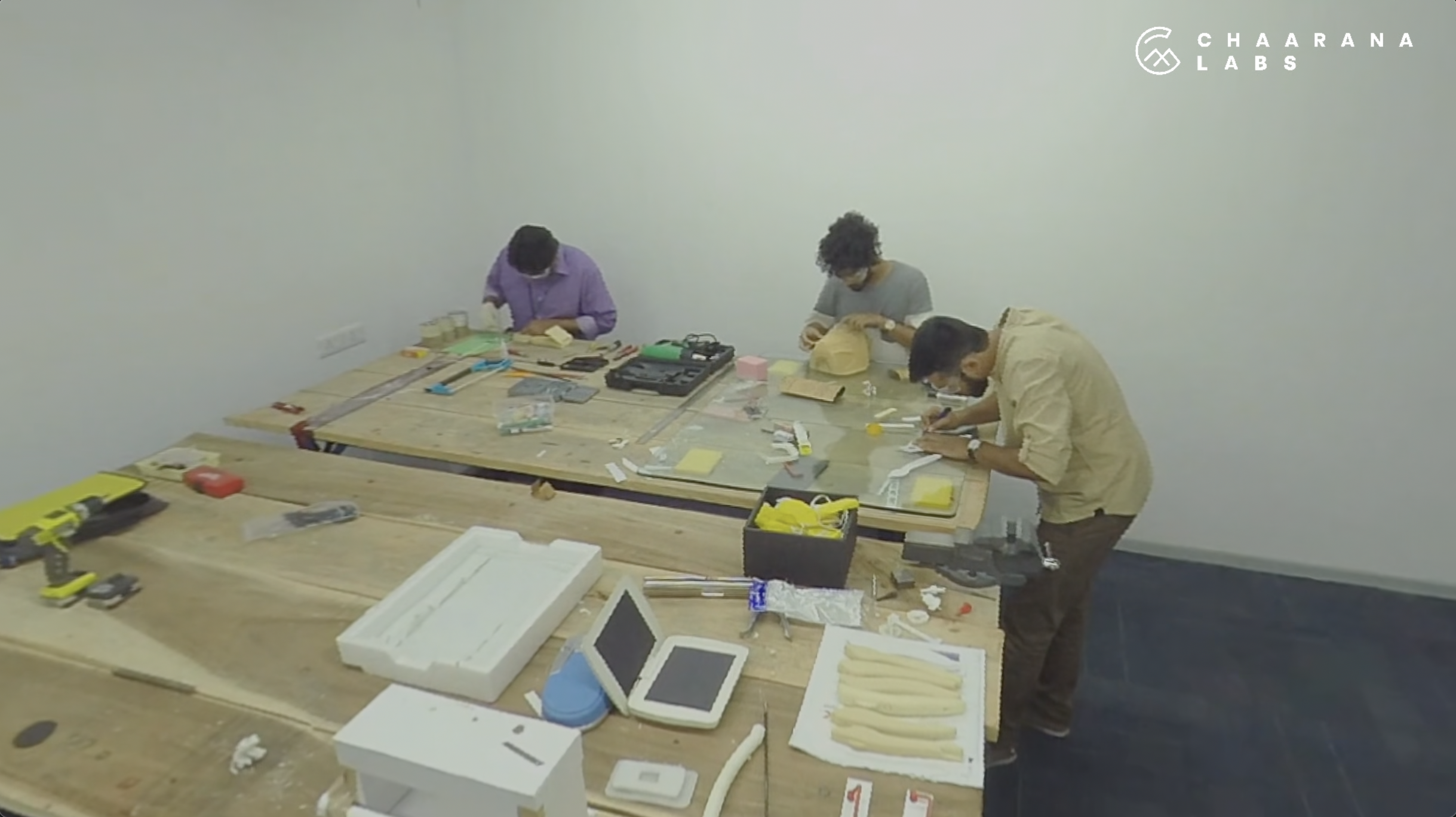 Three people working at a cluttered table with electronic components, tools, and foam pieces in a workshop or lab environment, with 'Chaarana Labs' logo on the wall.
