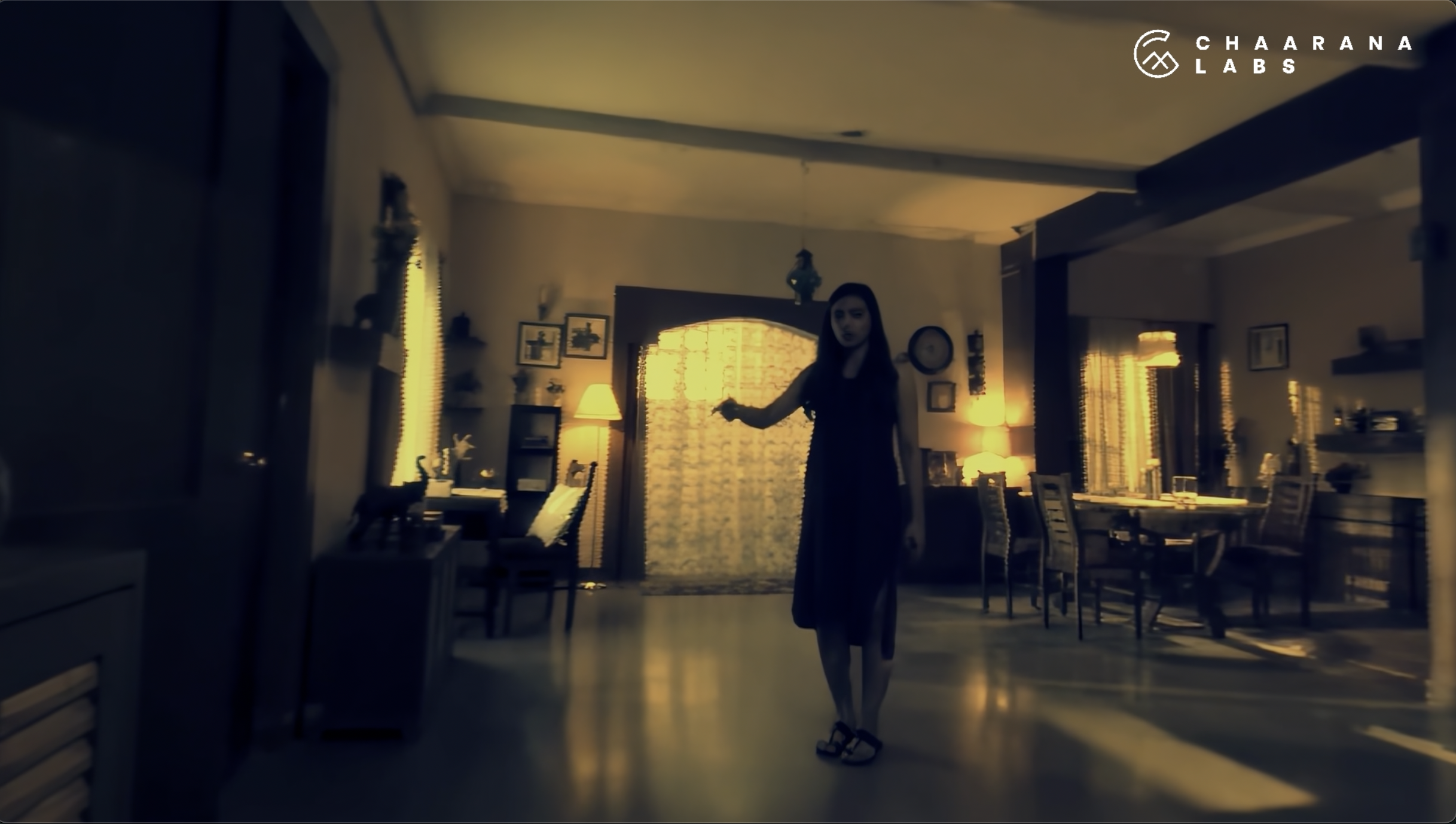 A woman in a long dress standing in a dimly lit dining room with yellow lighting, surrounded by chairs, tables, lamps, and picture frames on the walls. The room has a cozy, vintage atmosphere.