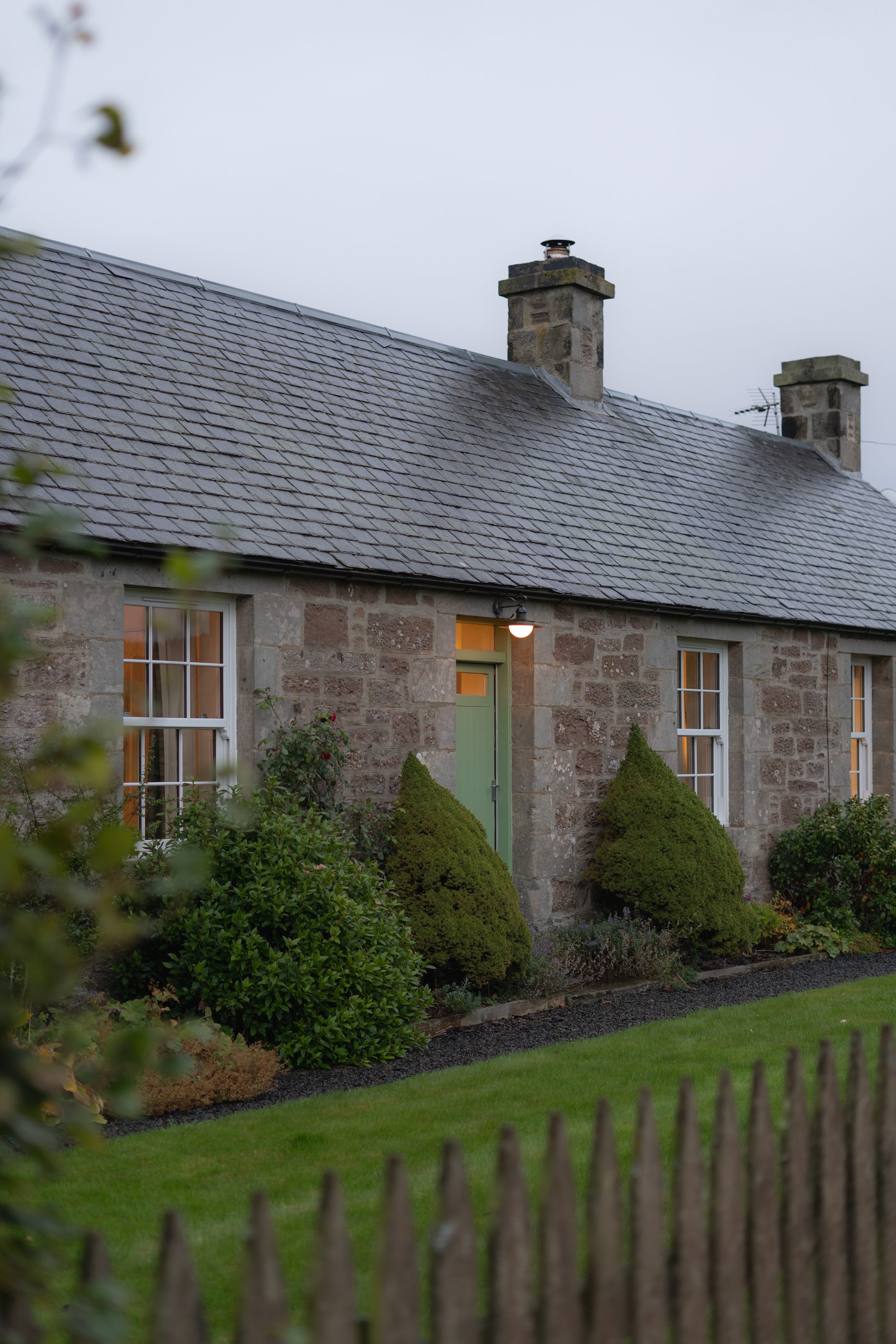 Stone cottage with green door and windows, surrounded by trimmed bushes and green lawn; overcast sky.