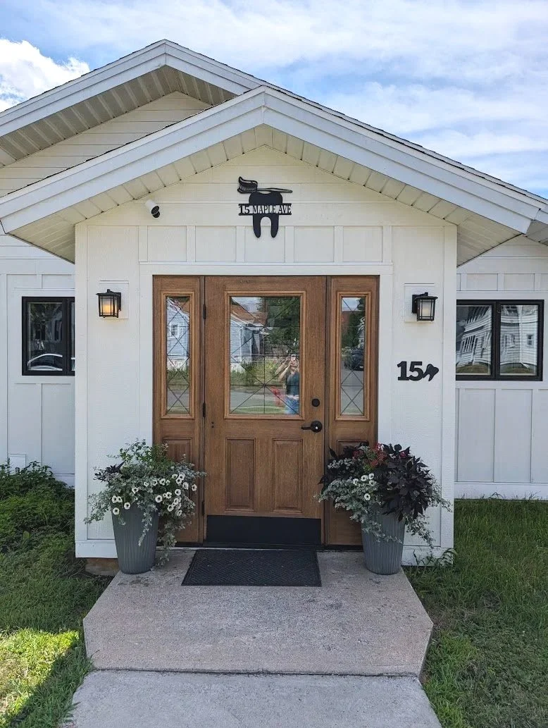 Front entrance of a white house with a wooden door, black lantern lights, and decorative signs including a black cat silhouette and the number 15. Potted plants with flowers are on either side of the door.