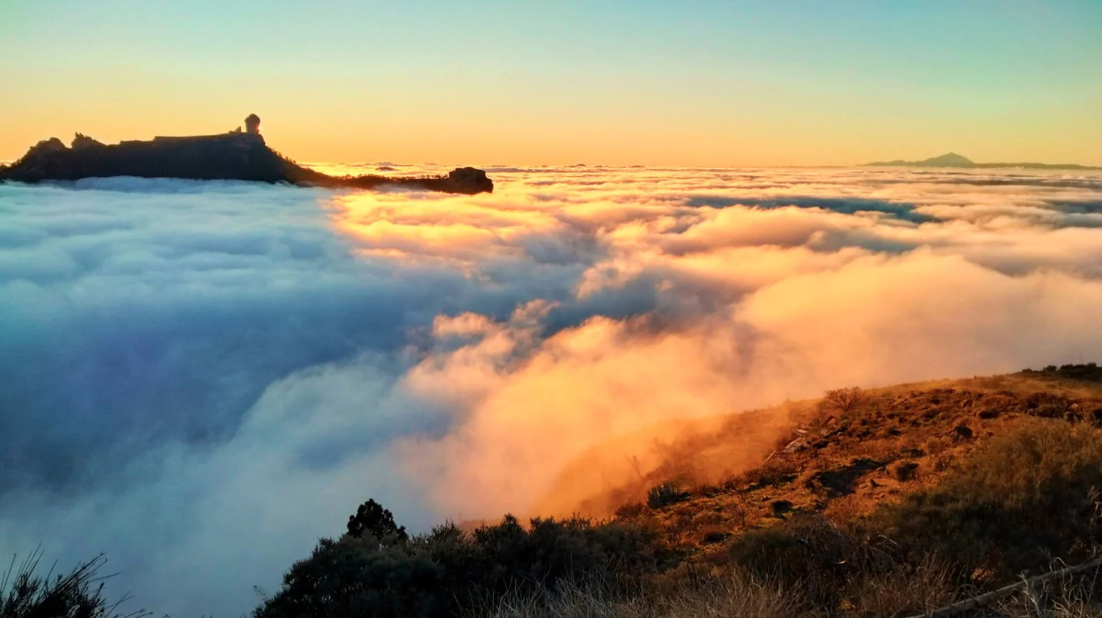 Sunset over a mountain covered in clouds, with a building or structure on top, and a hillside in the foreground.