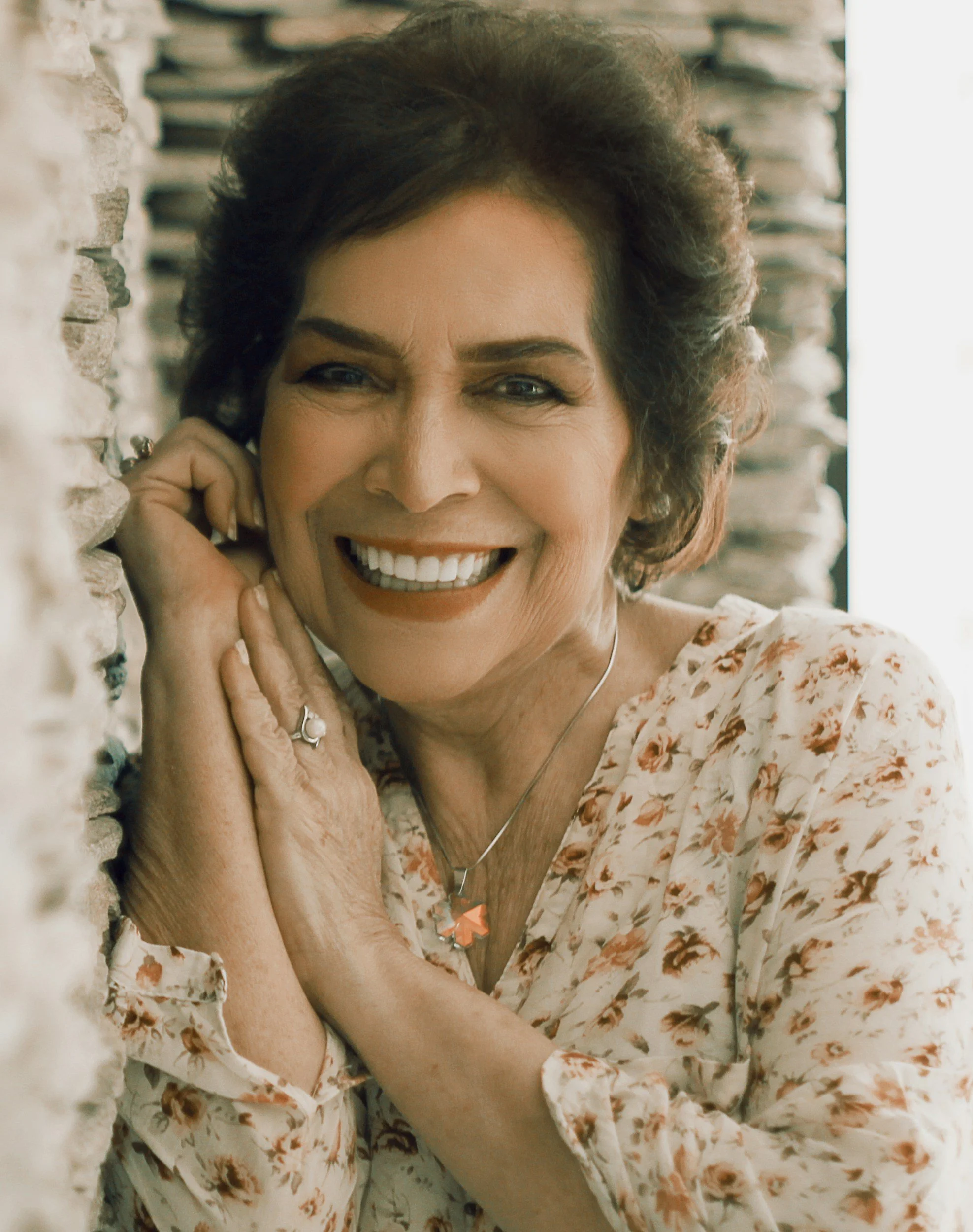 woman in a flower patterned shirt resting her hands by her face smiling