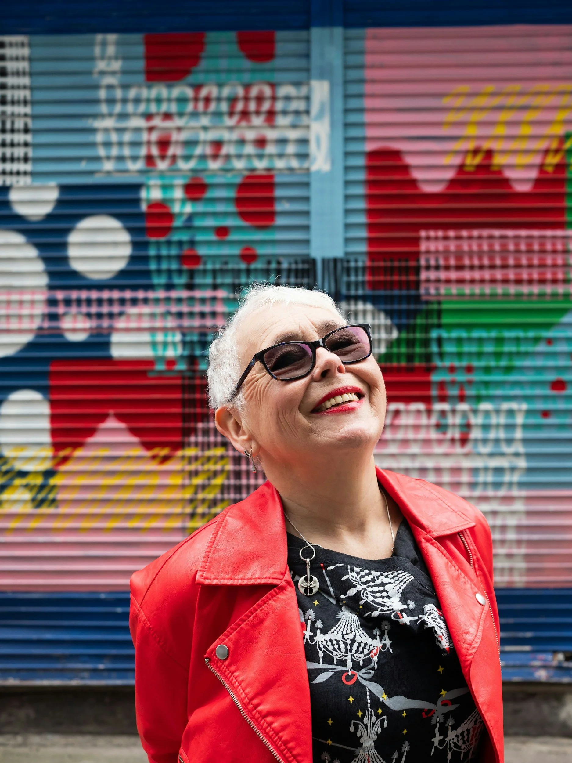 woman with white hair and black glasses wearing a red jacket standing and smiling in front of a mural