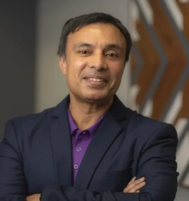 headshot of a man smiling with his arms crossed wearing a purple shirt with a black jacket