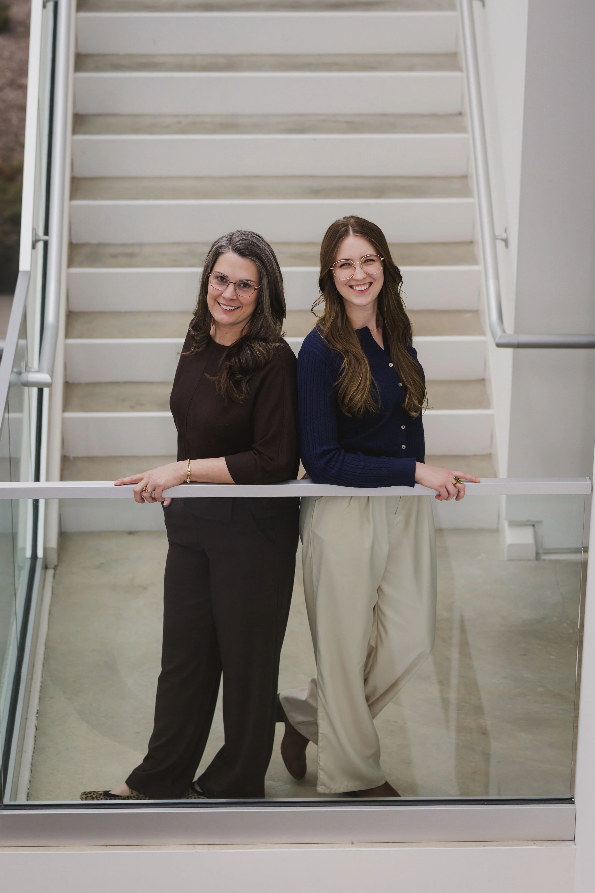 Heidi and Ella standing by a railing smiling both wearing glasses and dark shirts
