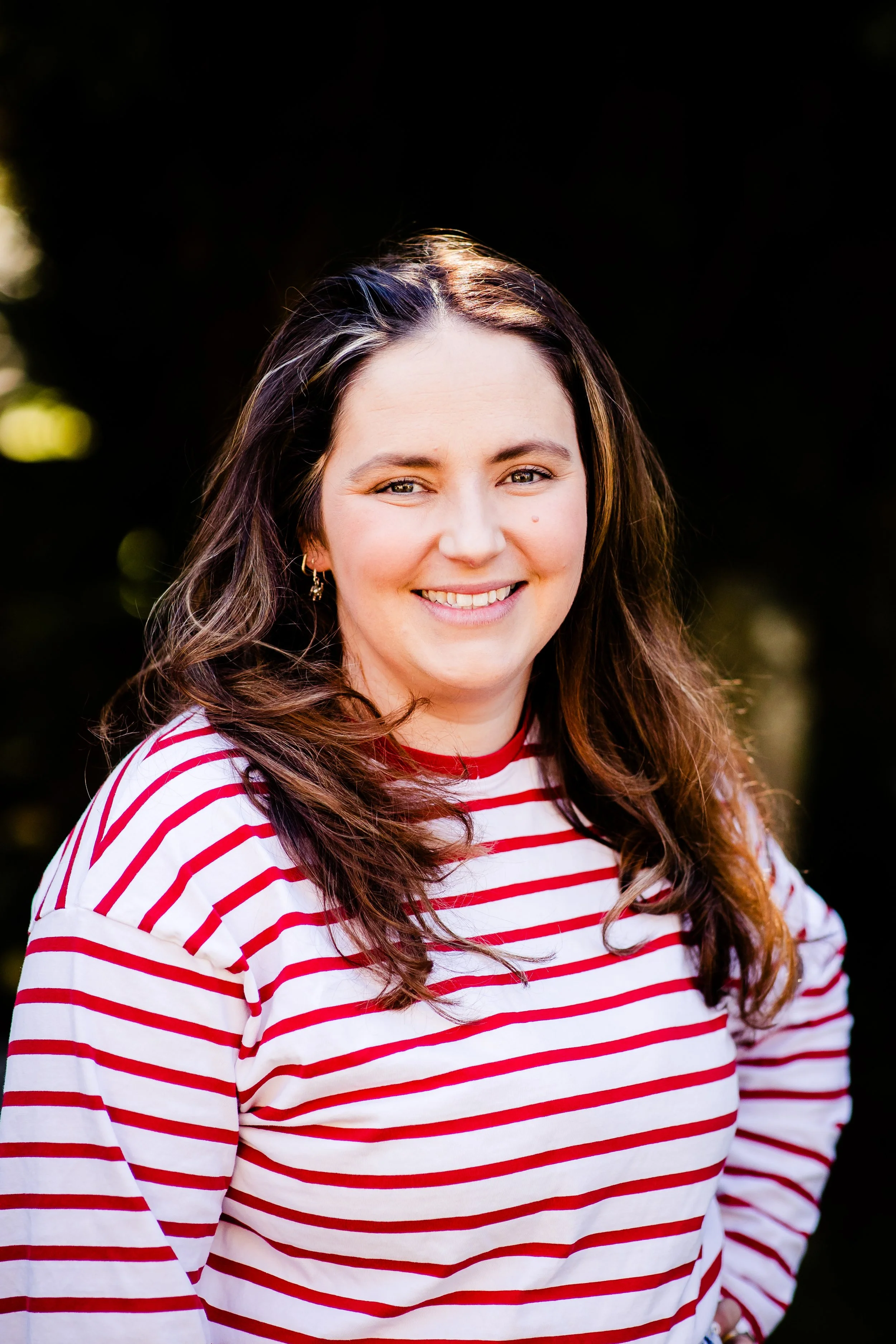 A young woman with long, wavy brown hair, wearing a white and red striped shirt, smiling outdoors against a dark background with blurred greenery.